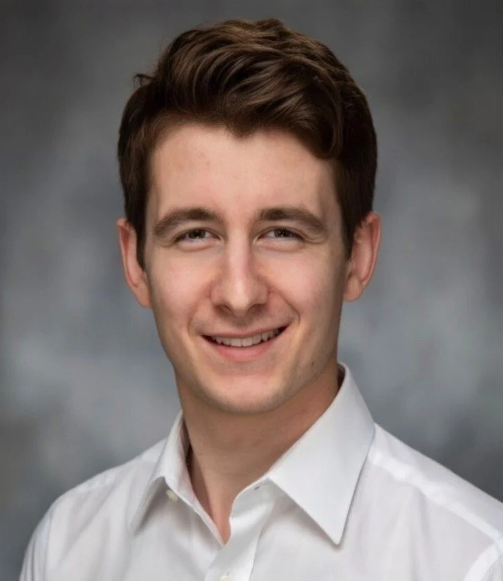 A young man with short brown hair, wearing a white collared shirt, smiling against a gray background.