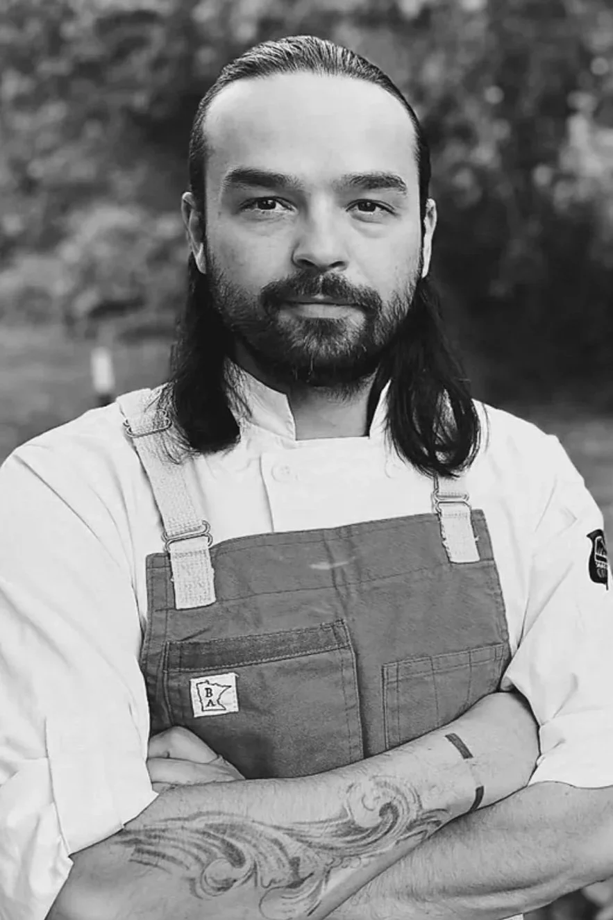 Black and white portrait of a chef with long dark hair, beard, wearing a chef's coat and apron, standing outdoors with arms crossed, background of trees.