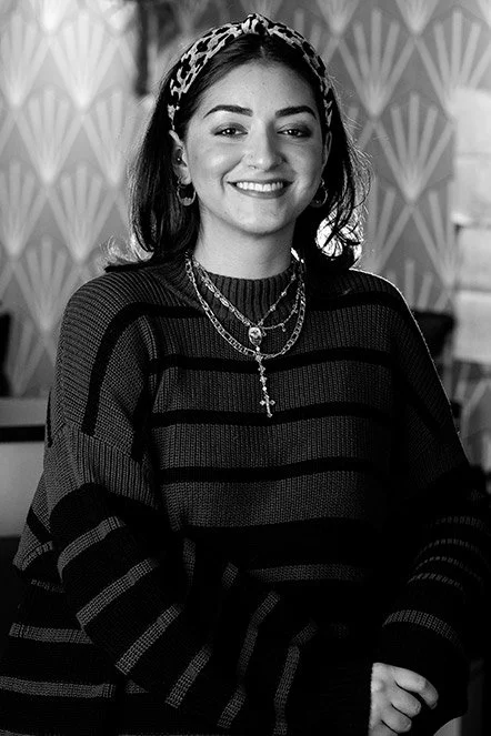 Black and white photo of a young woman with dark hair, wearing a patterned headband, layered necklaces, earrings, and a striped sweater, smiling at the camera indoors.