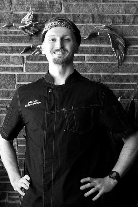 A smiling man wearing a chef's coat and a headband, standing in front of a brick wall with decorative fish sculptures.