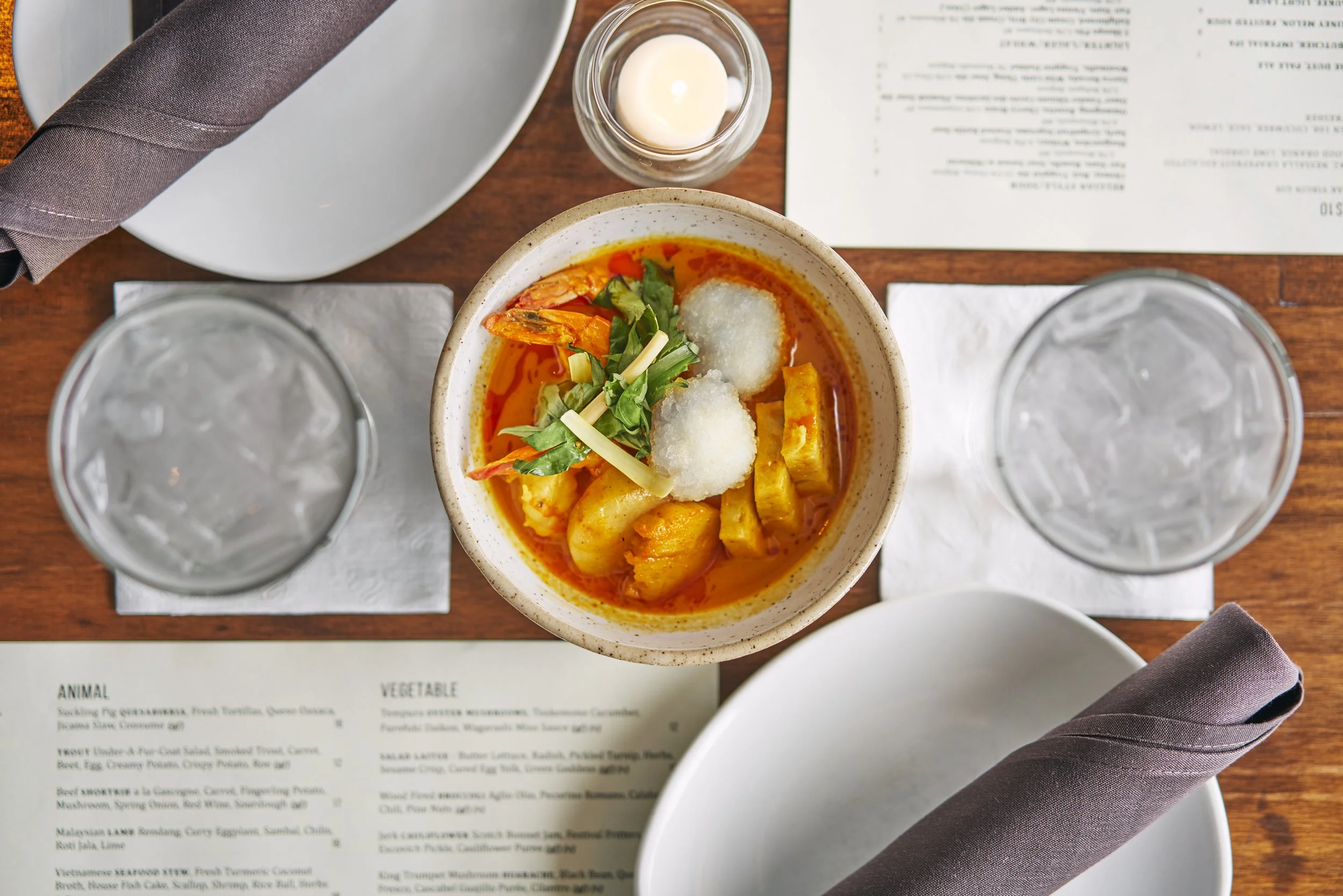 A bowl of spicy seafood stew garnished with green herbs on a wooden table, accompanied by a glass of water, a candle, a plate with a folded napkin, and a menu.
