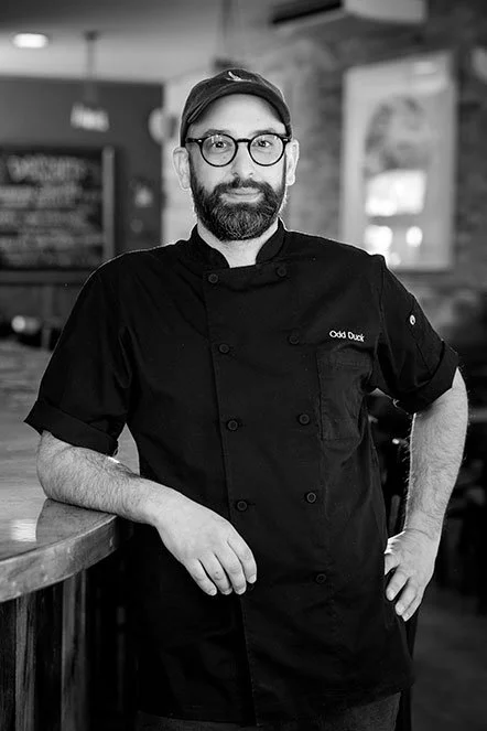 A man in a chef's uniform with glasses and a cap, standing in a restaurant kitchen.