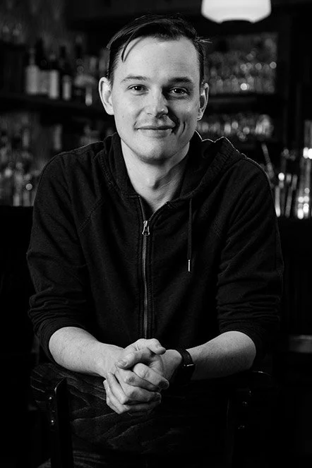 A young man with dark hair and light skin sitting at a bar counter, smiling, with bottles and glasses in the background.