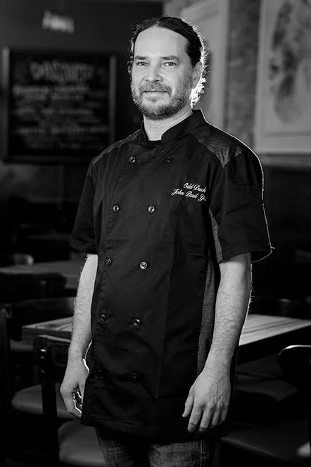 Black and white photo of a male chef in a kitchen, wearing a chef's coat with embroidered text.