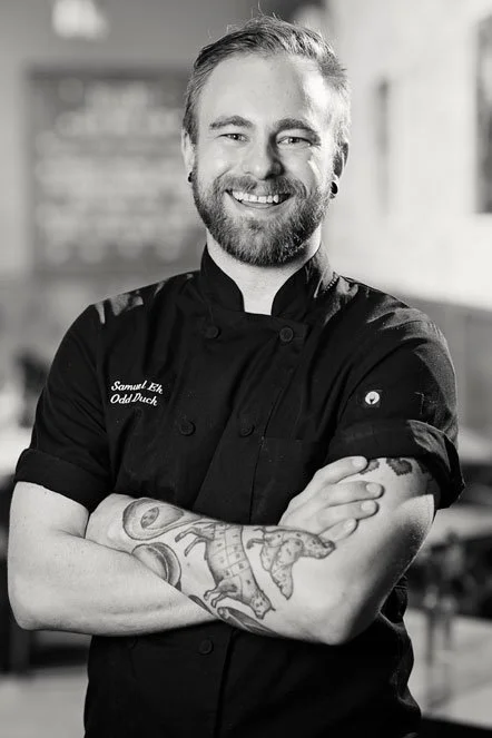 Portrait of a smiling male chef with a beard, wearing a black chef's coat with his arms crossed, tattoos on his arms, in a dining setting.