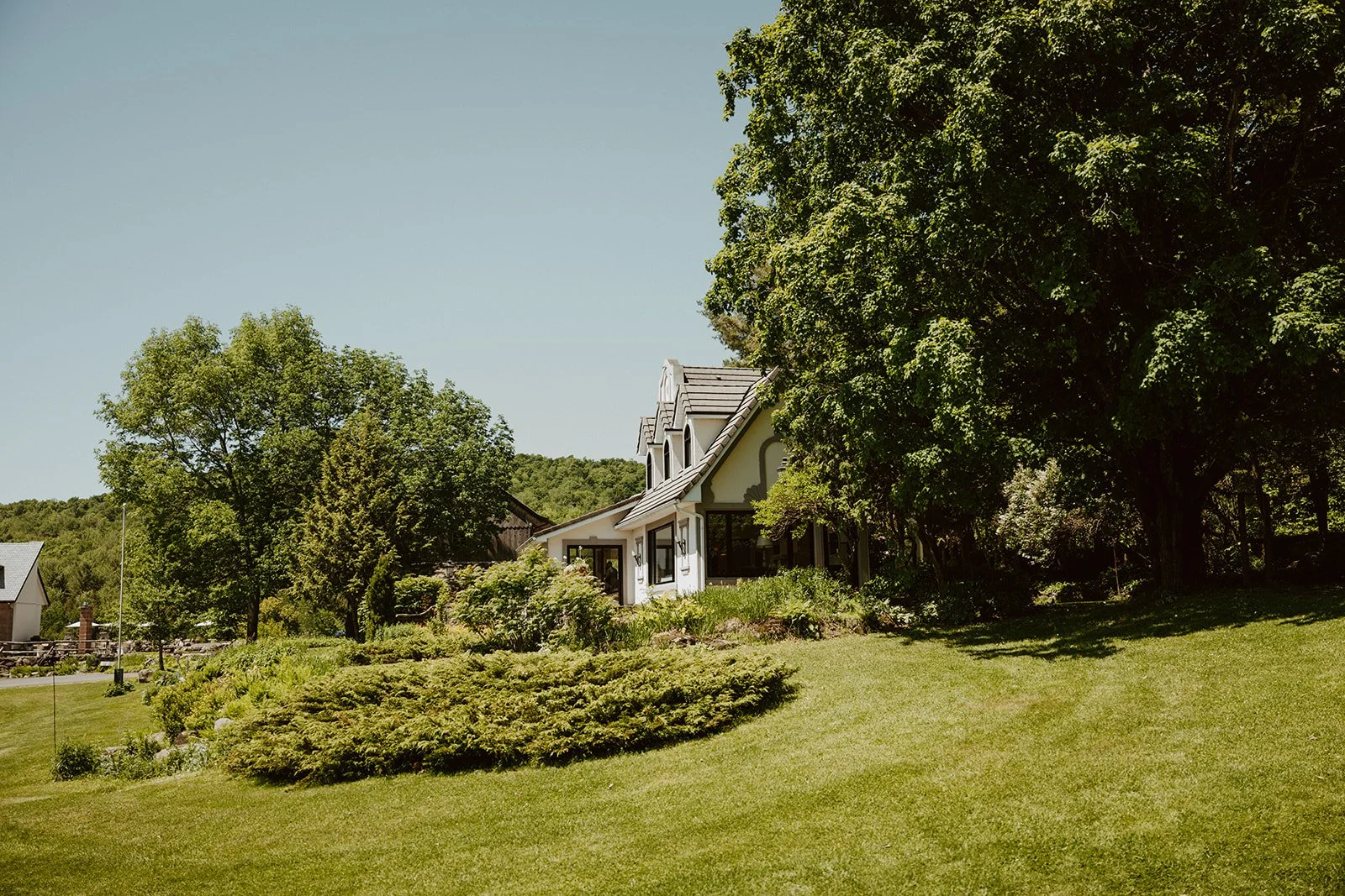 A house with multiple gabled roof sections is surrounded by lush green trees and a well-maintained lawn, with shrubs and plants in the foreground, under a clear blue sky.