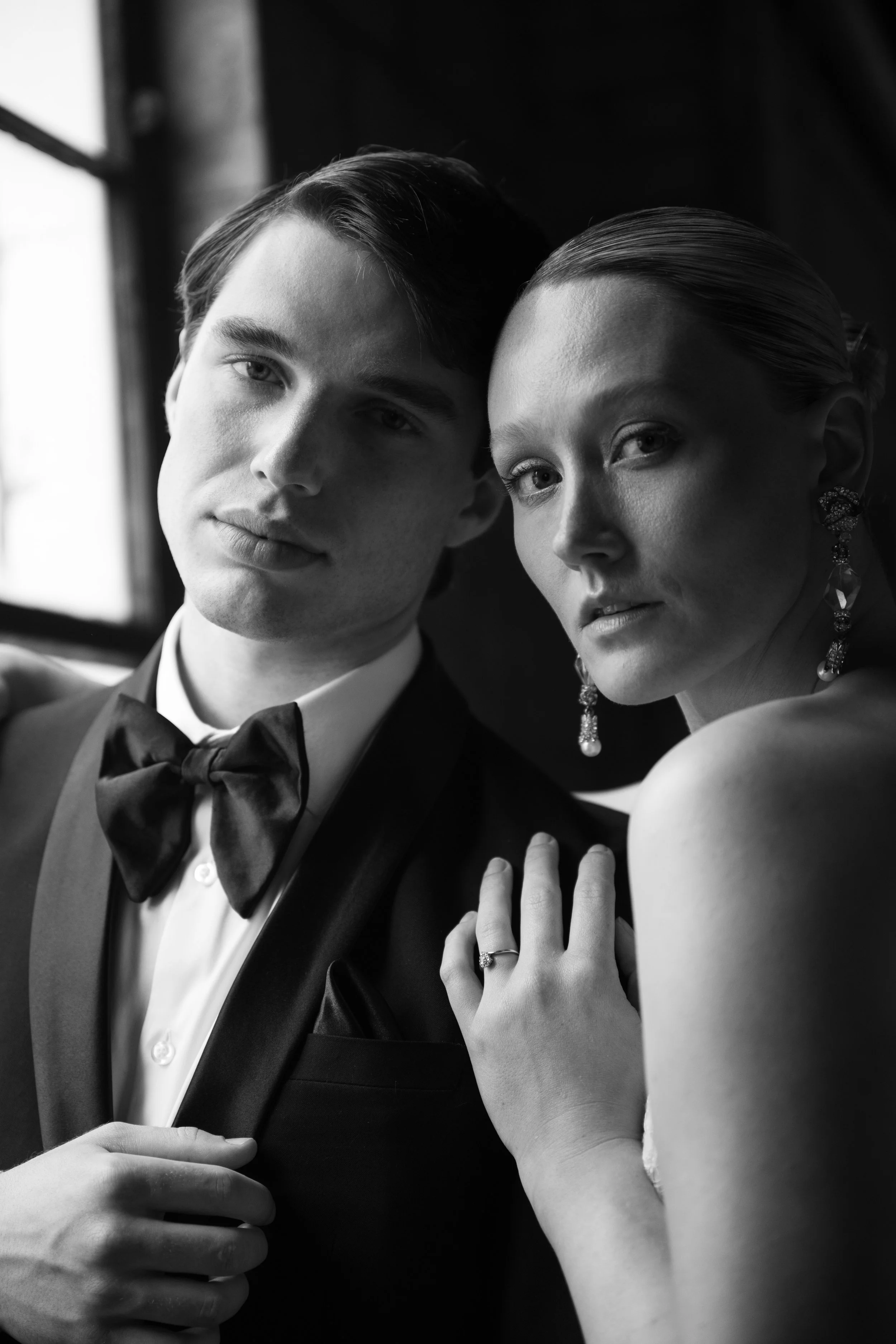 Black and white photo of a man in a tuxedo with a bow tie and a woman with elegant earrings, posing closely together near a window.