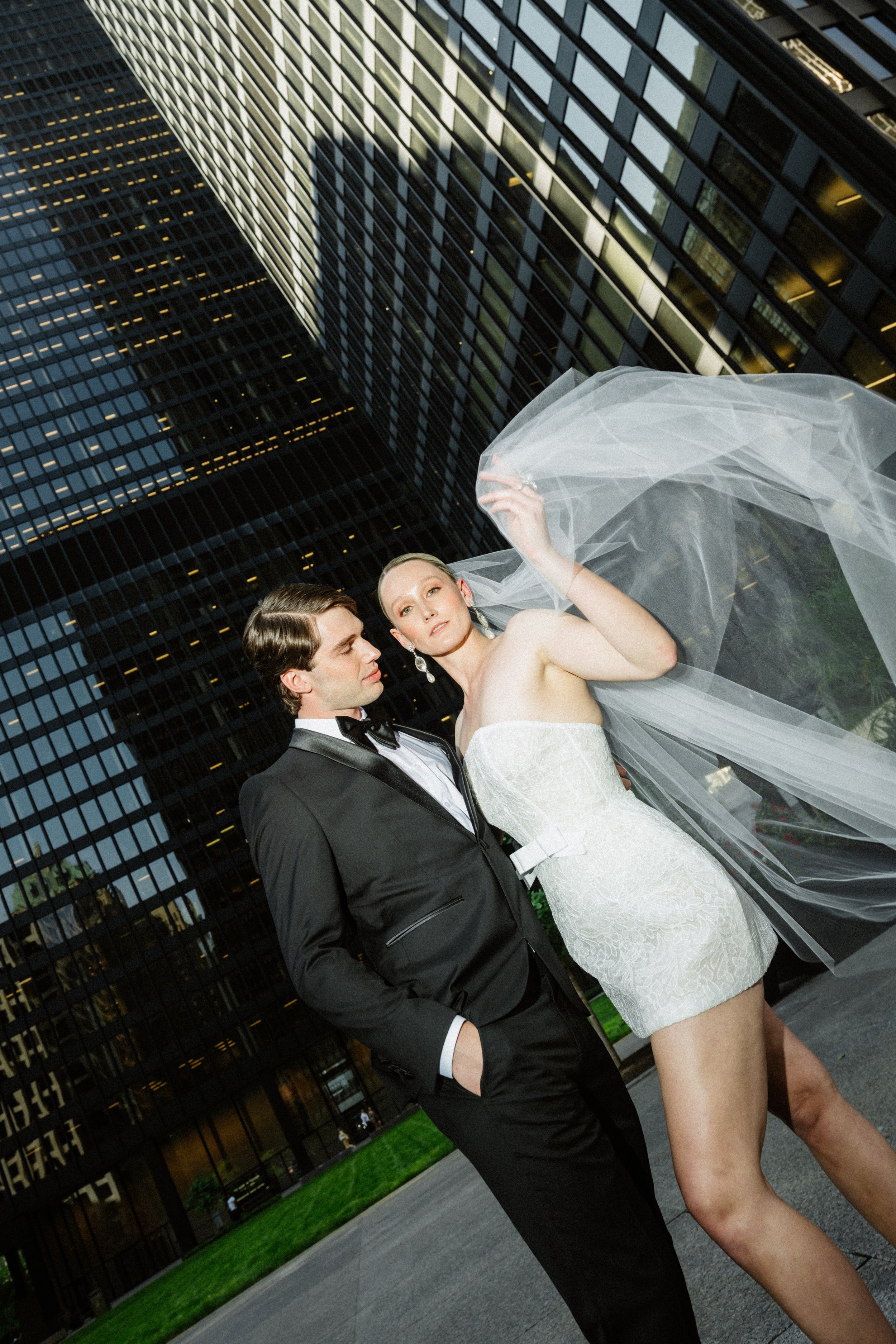 A fashionable couple in wedding attire standing in front of tall modern glass buildings during the evening.