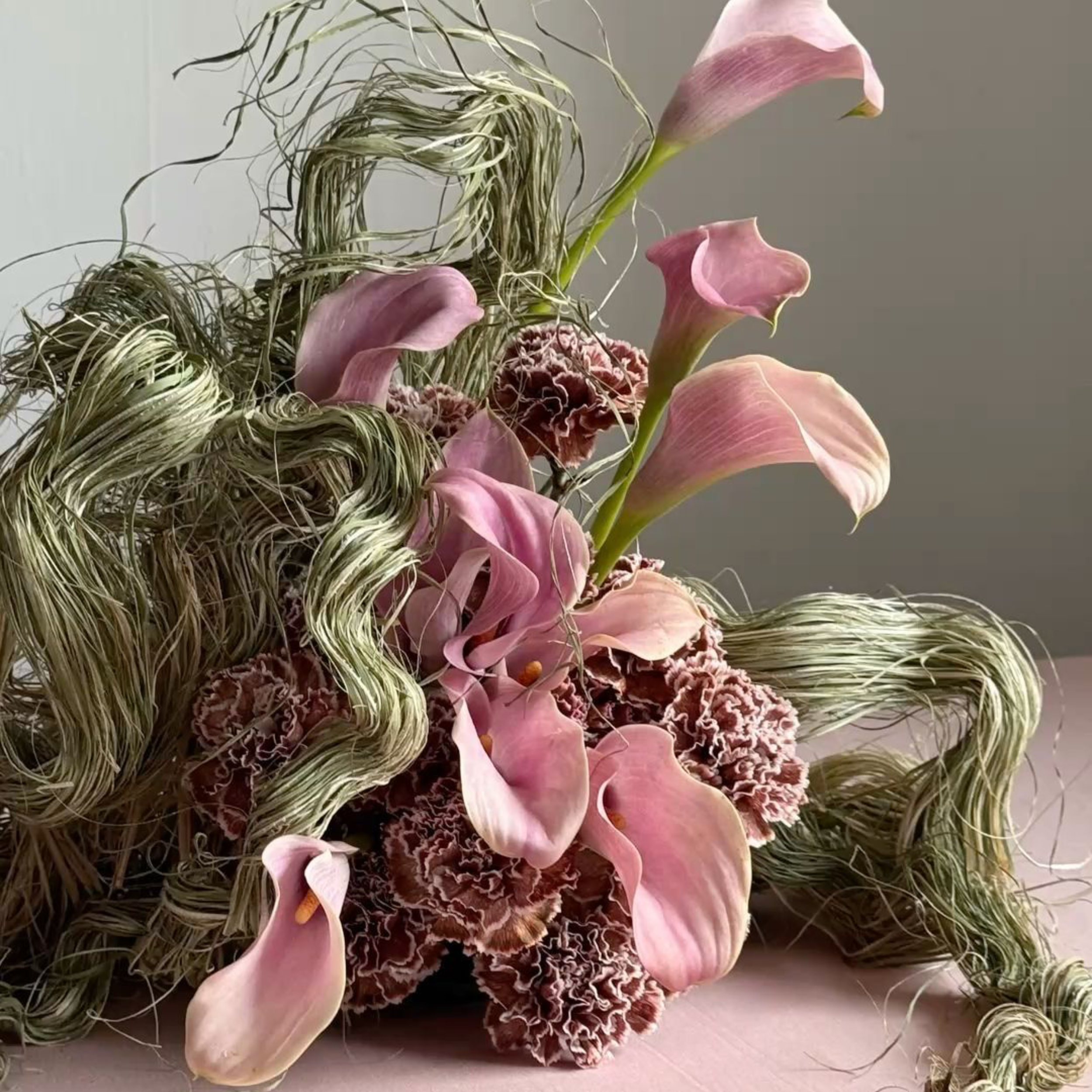 A floral arrangement with pink calla lilies, ruffled brown and pink flowers, and curly green and cream-colored foliage.