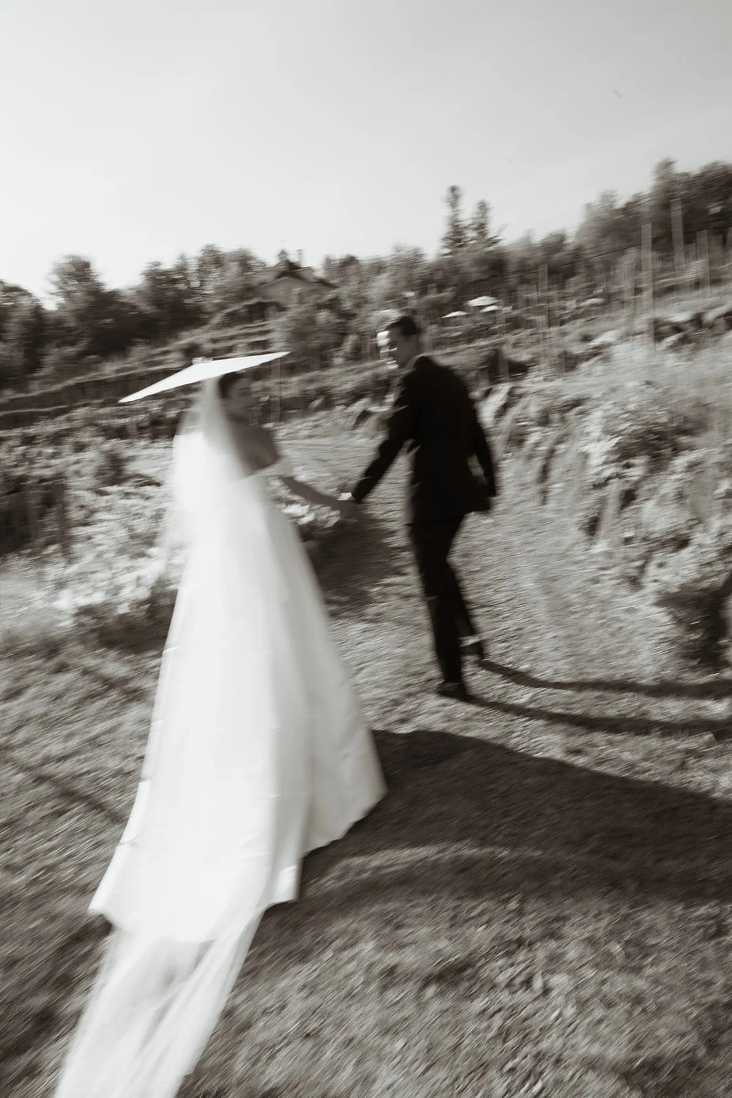 A blurred black and white photo of a bride in a long wedding dress holding hands with a groom in a suit, walking along a dirt path on a hillside.