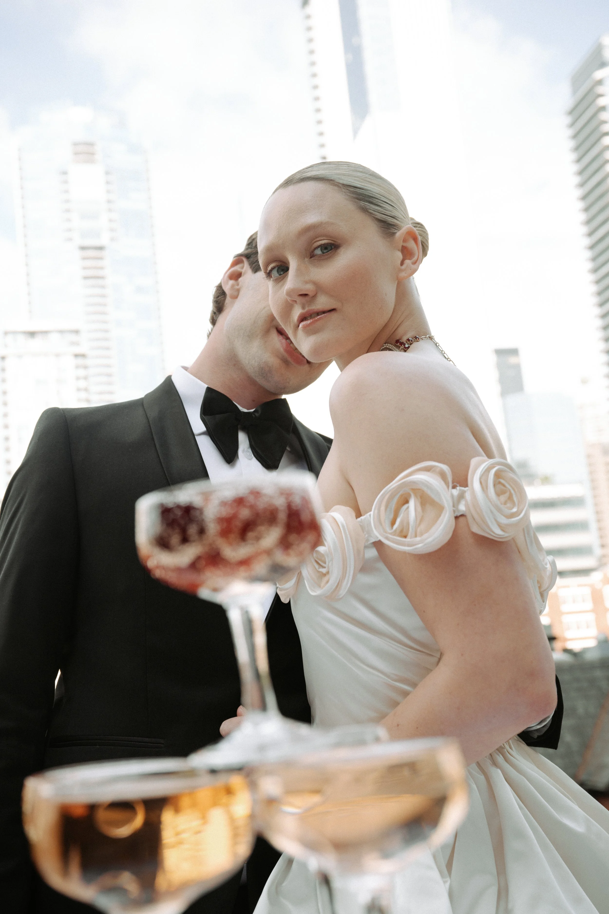 A couple dressed in formal attire on a city rooftop, with wine glasses in front of them, skyscrapers in the background.