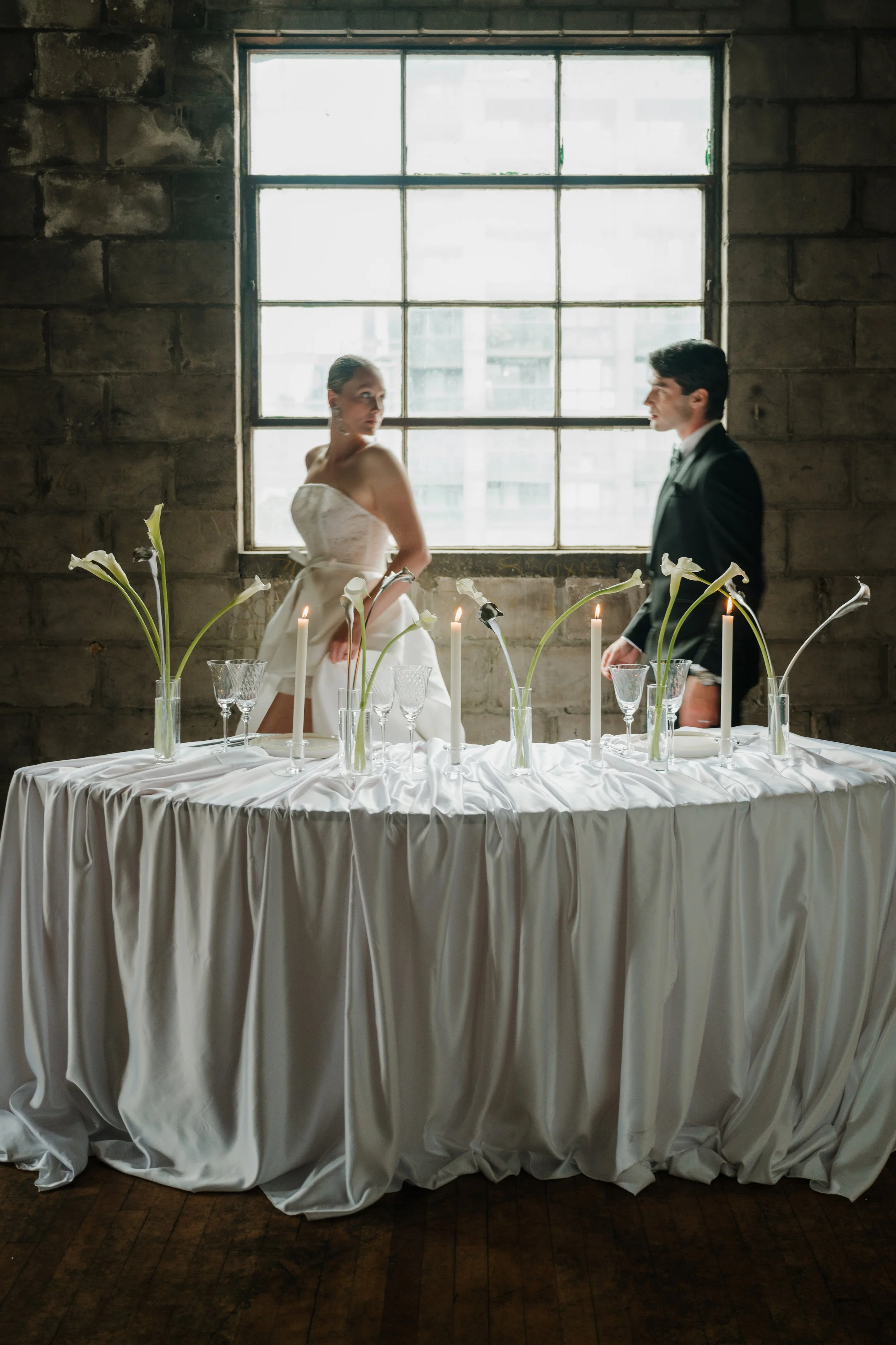 A bride and groom standing on opposite sides of a table with a white tablecloth, decorated with calla lilies, candles, and glassware, in front of a large window with a brick wall behind them.