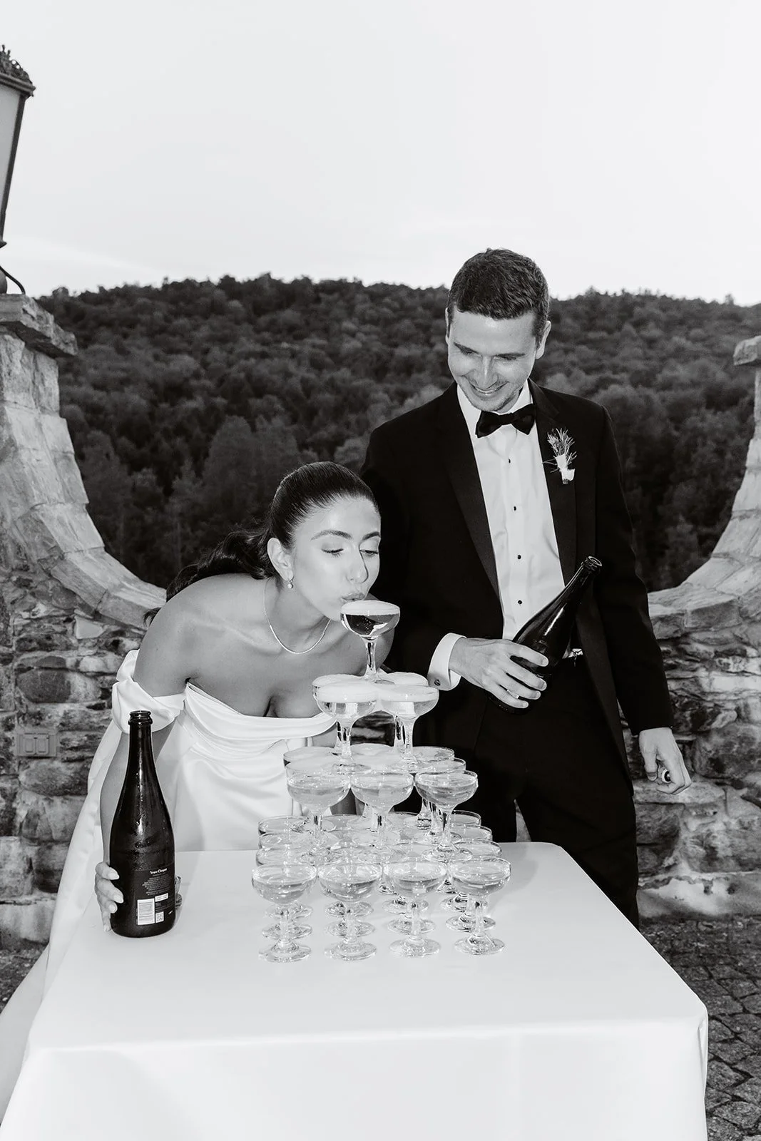 Bride and groom participating in a champagne tower toast at their wedding, outdoors with a scenic background of trees and mountains.