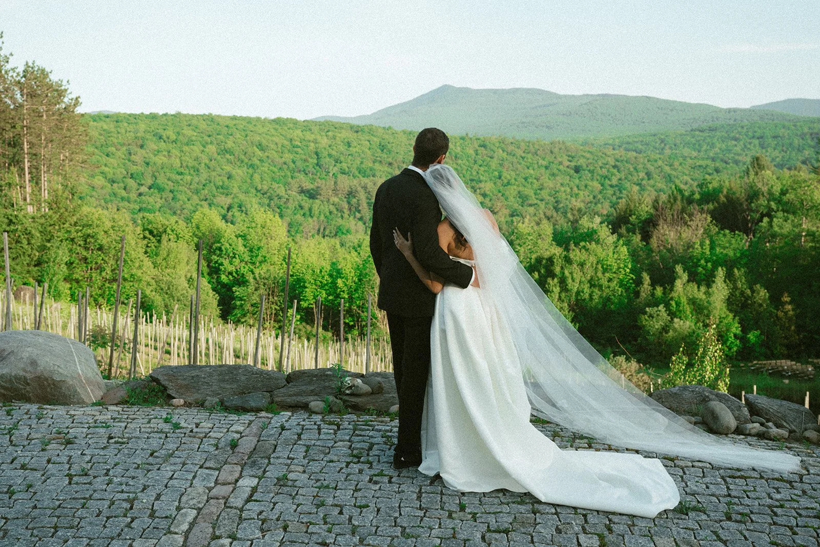 Bride and groom embracing outdoors on cobblestone area with green forest and mountains in background.