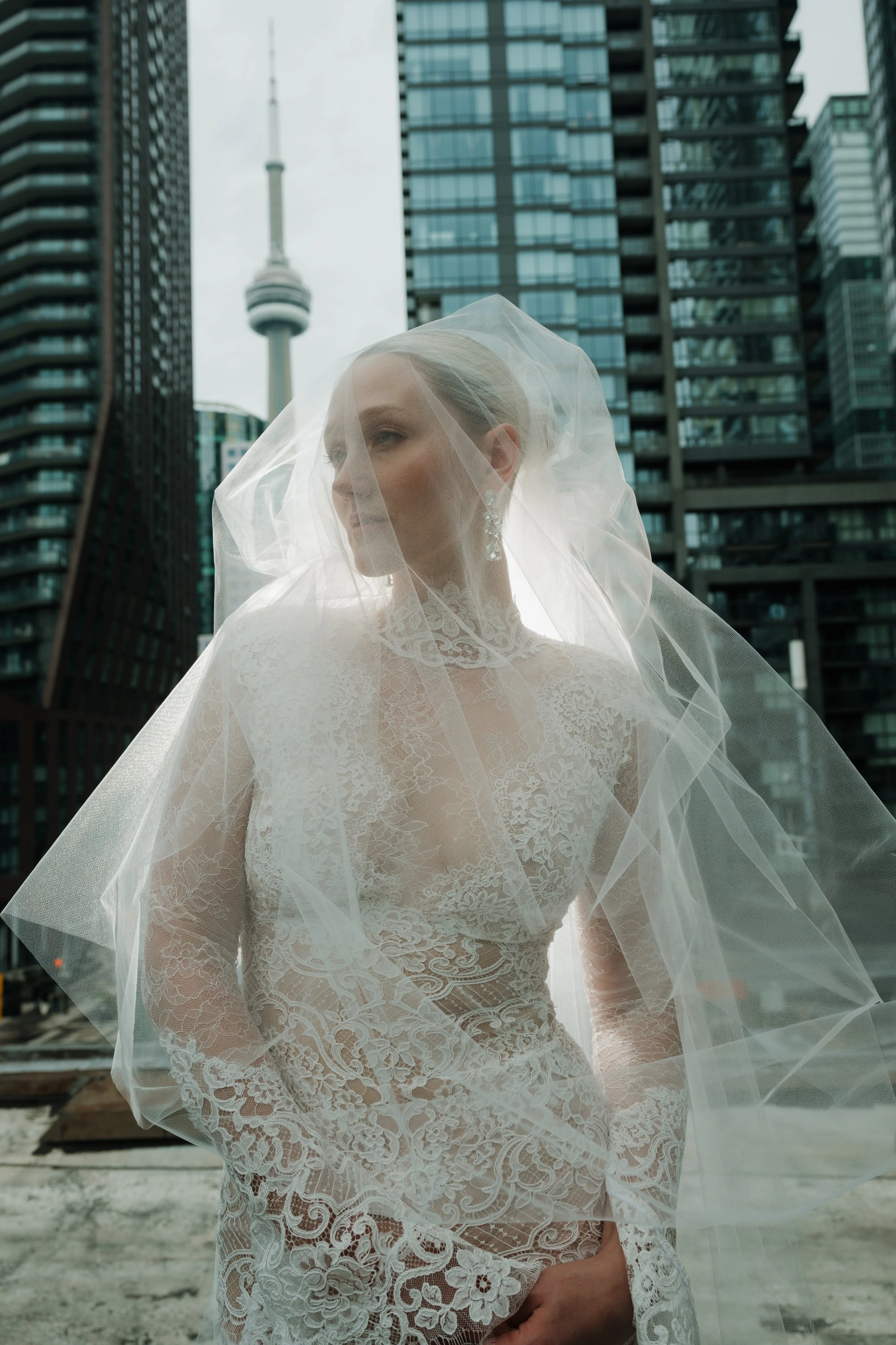 A bride in a lace wedding dress and veil standing outdoors in an urban setting with modern skyscrapers and a tall TV tower in the background.