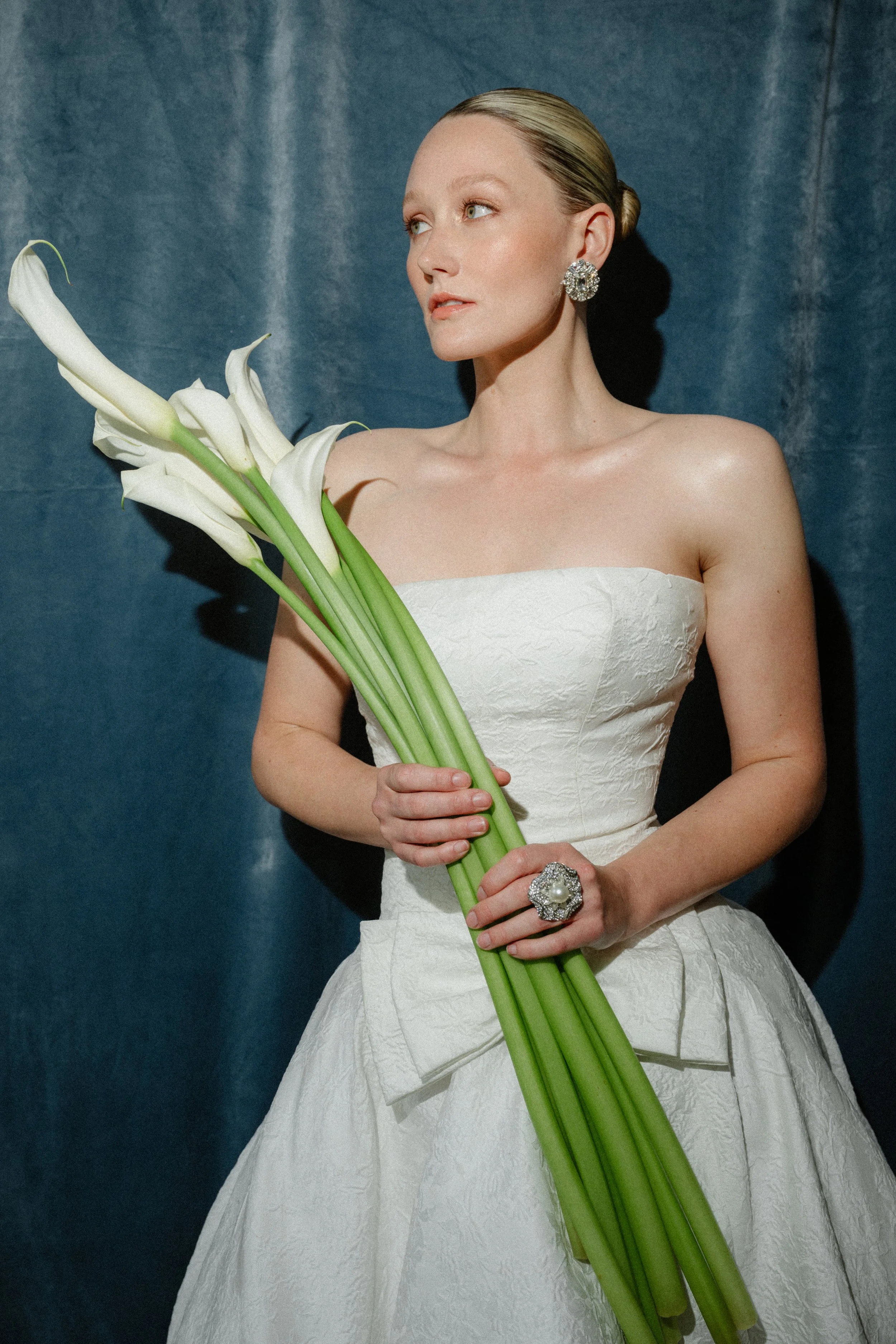 A woman in a white strapless dress holding white calla lilies, standing against a blue background.