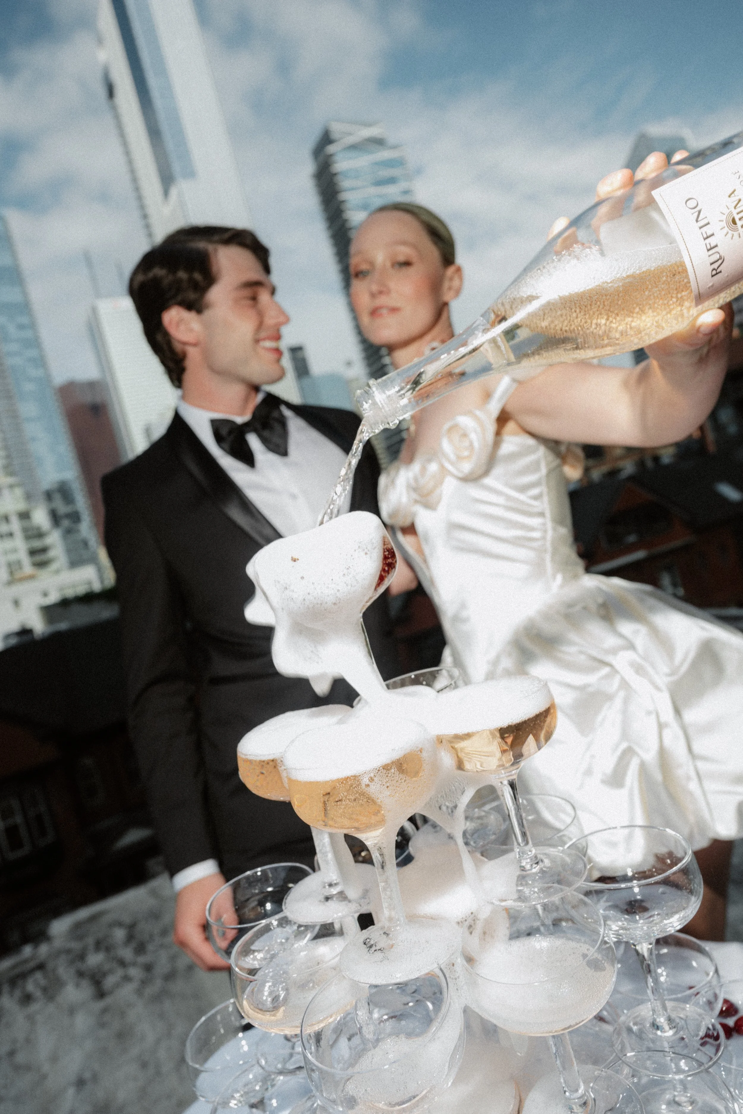 A man and woman in formal attire, with the man in a tuxedo and the woman in a white dress, are celebrating on a rooftop with a champagne tower, under a cloudy sky with high-rise buildings in the background.