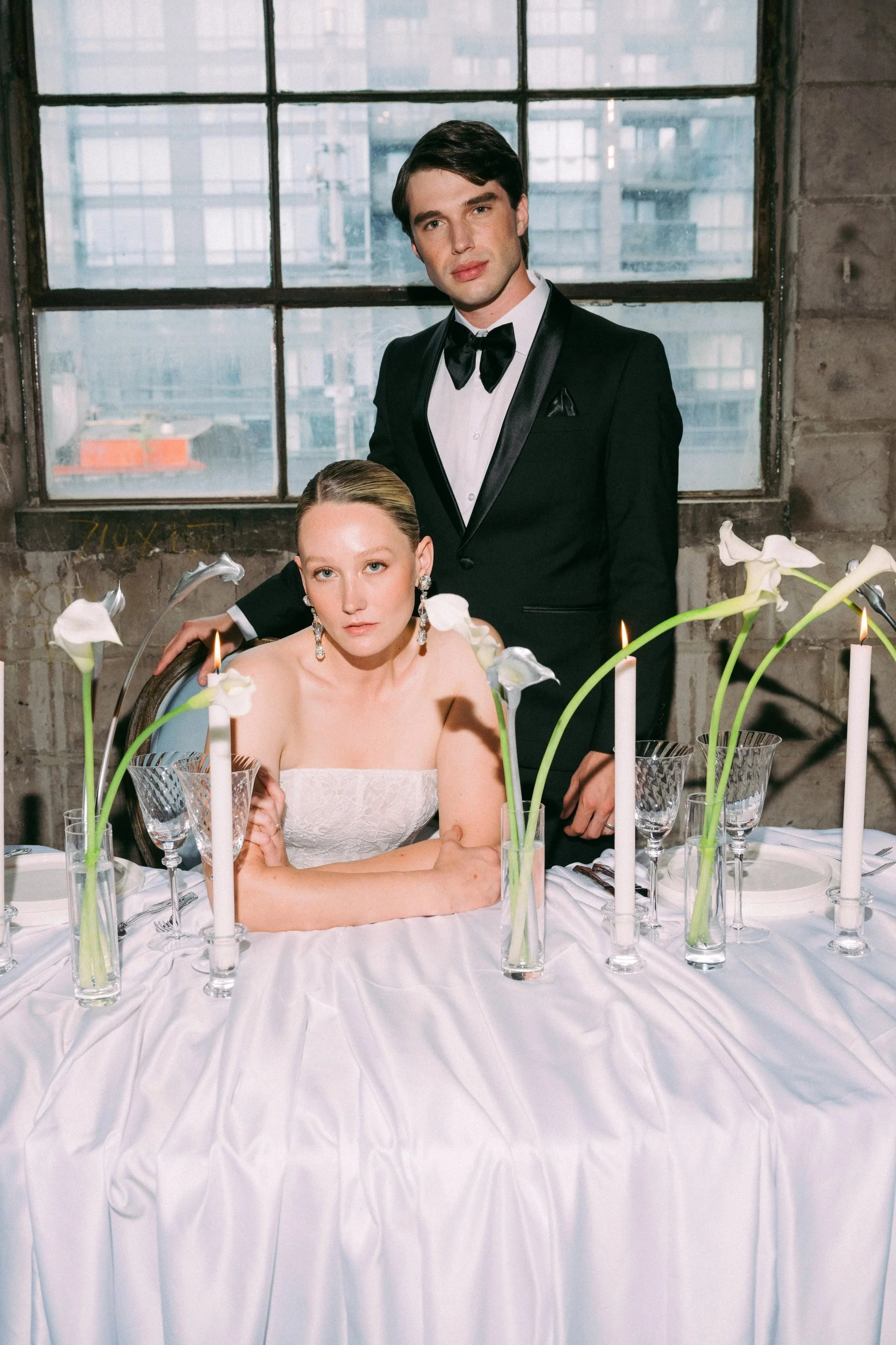 A man in a black tuxedo and a woman in a white strapless dress sit at a formal dinner table with white tablecloth, tall white candles, and calla lilies in glass vases. The woman is leaning on her arms, and the man stands behind her with one hand on her shoulder in an industrial-style room with large windows.