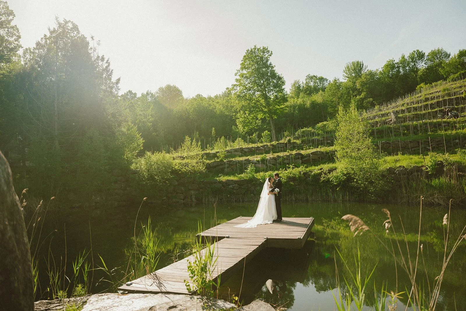 A bride and groom standing on a wooden dock on a small lake, embracing against a lush, green hillside with trees and terraced gardens, during sunset.