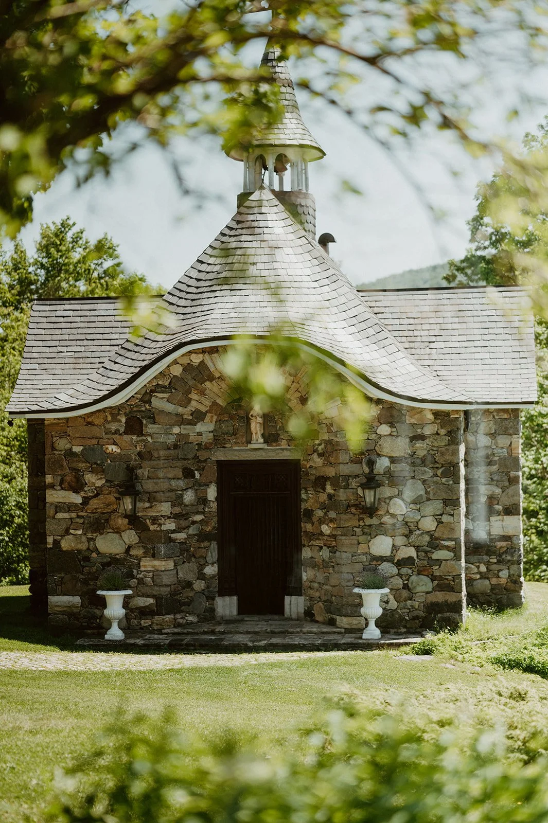 A small stone chapel with a curved roof and a small bell tower, surrounded by greenery and trees.