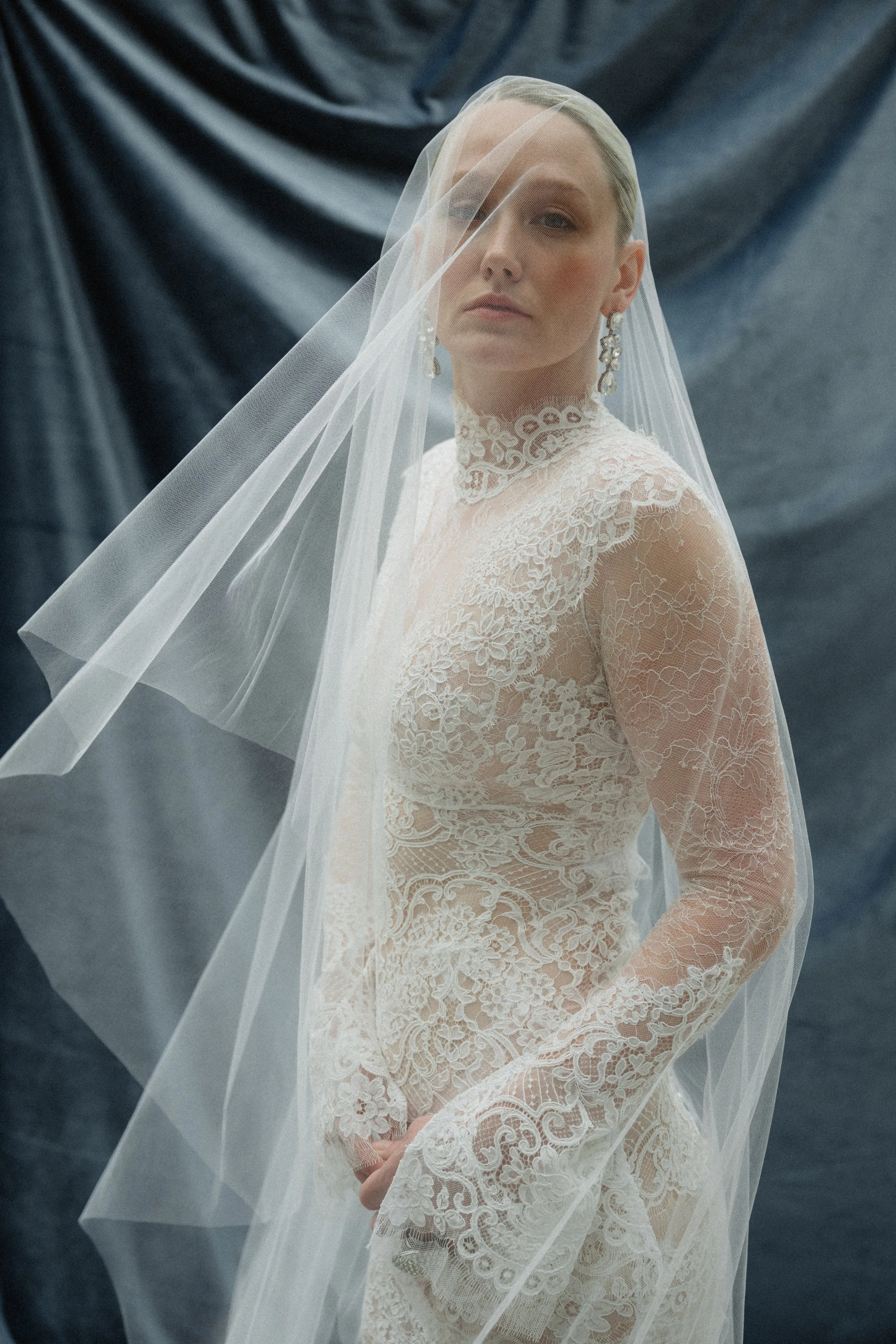 A woman in an elegant lace wedding dress with a veil over her face, posing against a dark fabric backdrop.