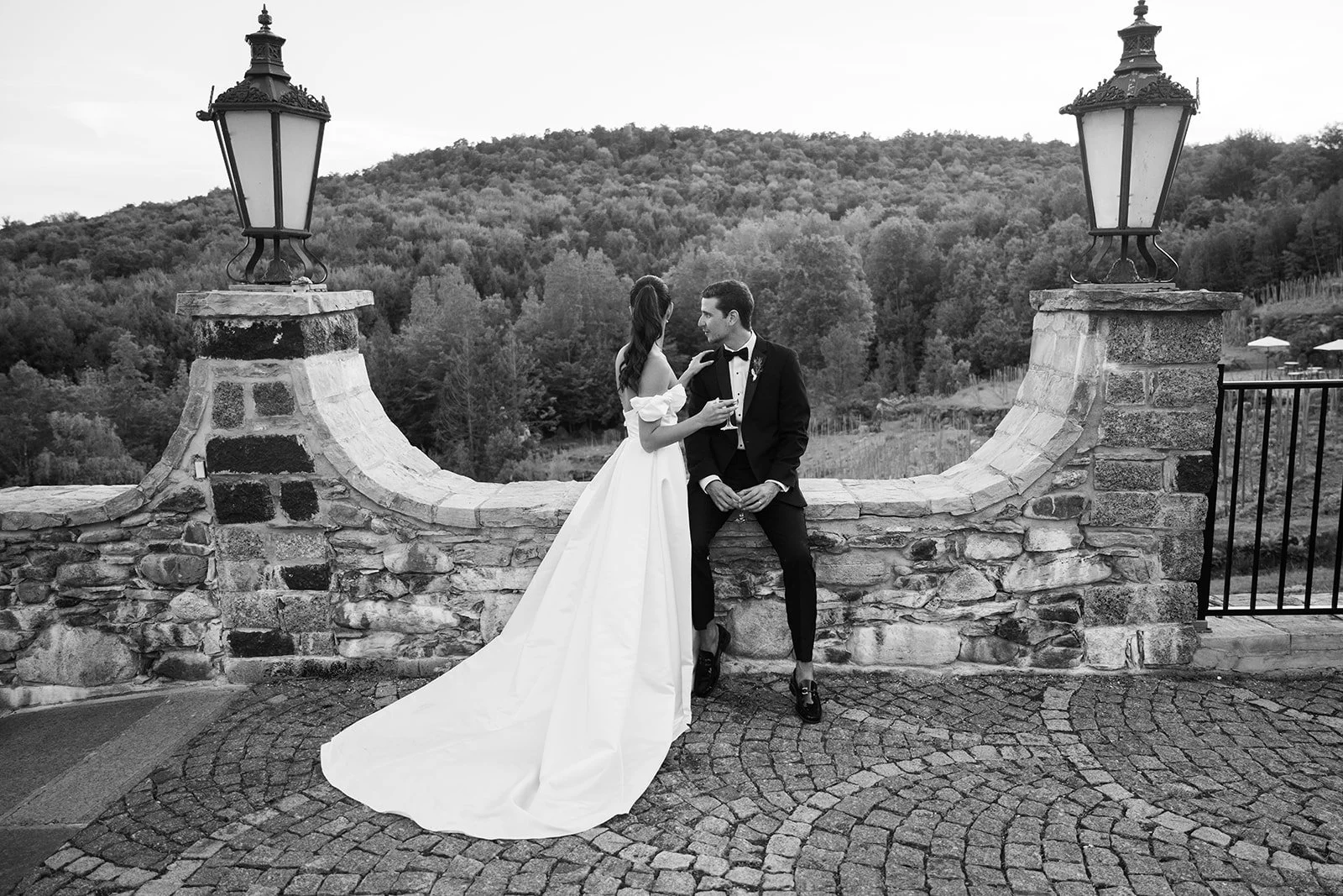 Black and white photo of a bride and groom on a stone terrace with scenic hilly background, the bride is in a wedding gown, and the groom is in a tuxedo.