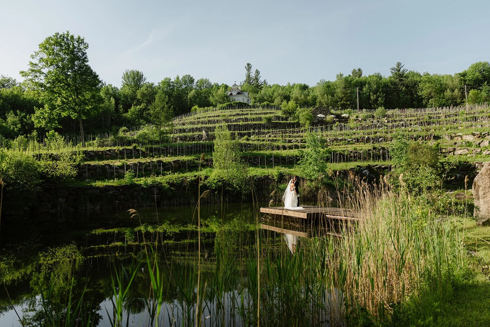A bride and groom standing on a wooden platform by a pond, with green trees and a terraced hillside in the background on a sunny day.