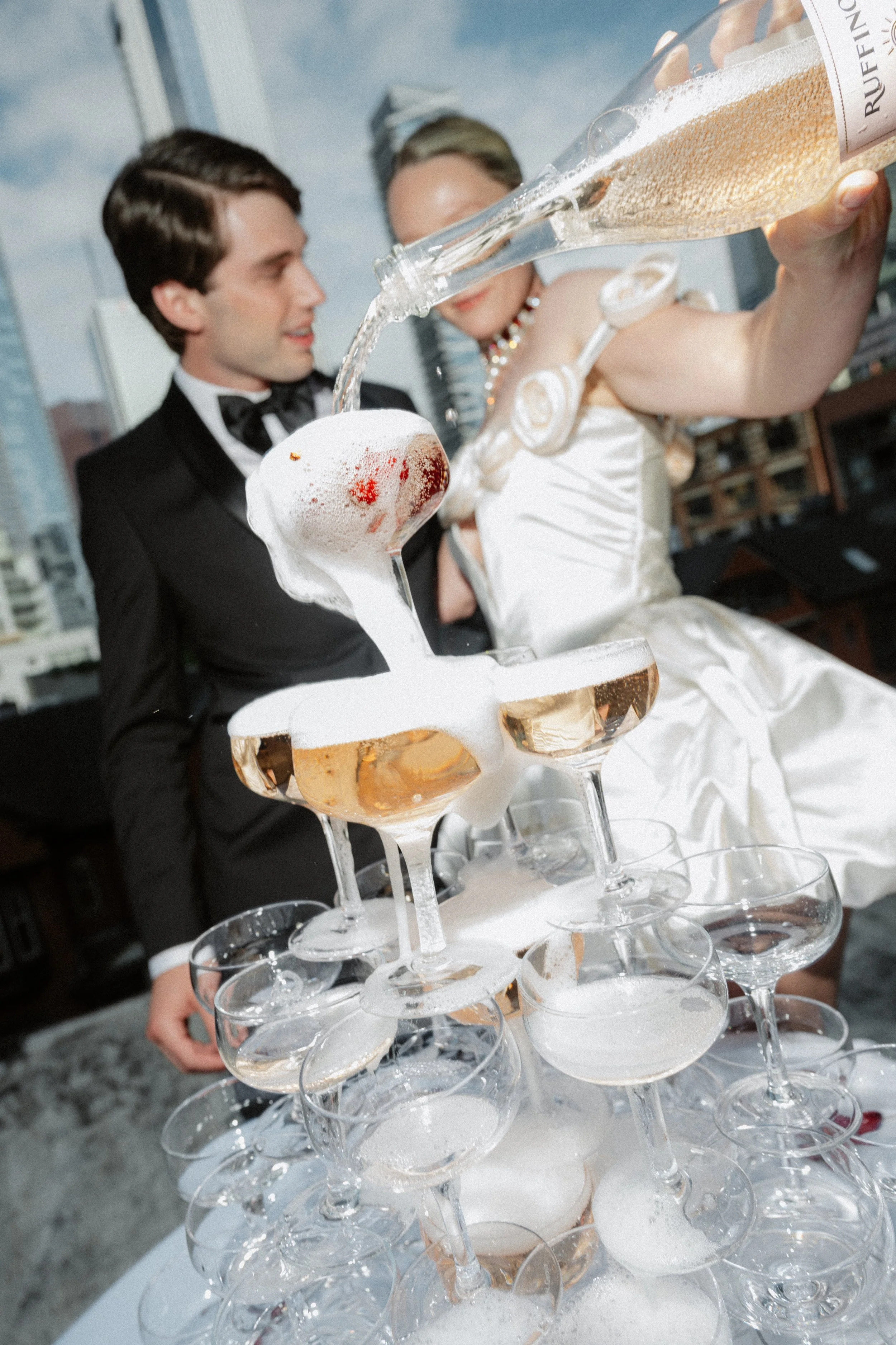 A man and woman in wedding attire pour champagne into a pyramid of champagne glasses, creating foam as the drinks overflow.