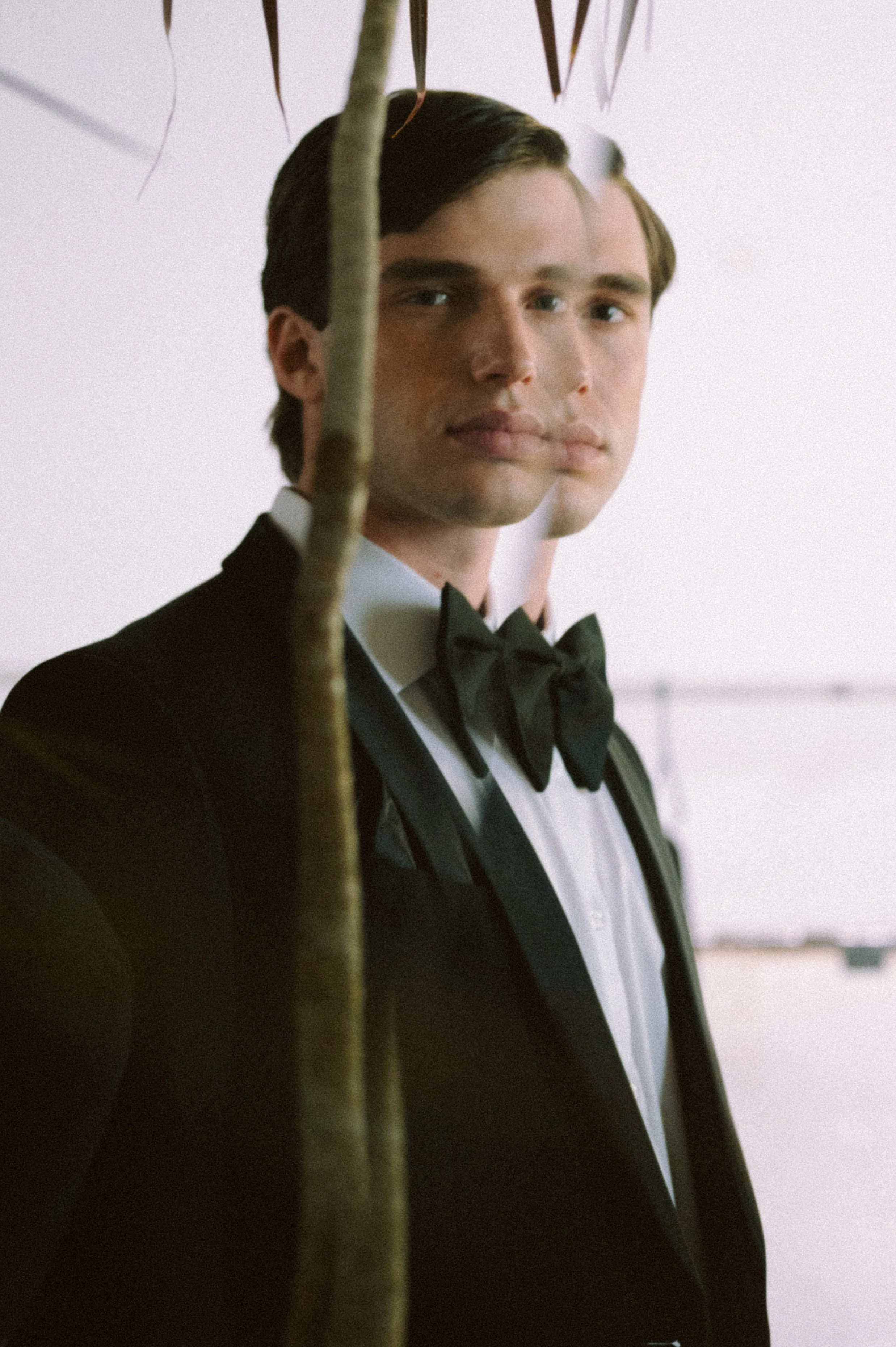 Young man in a tuxedo with a black bow tie, standing outdoors with a blurred background, partially obscured by a tree branch.