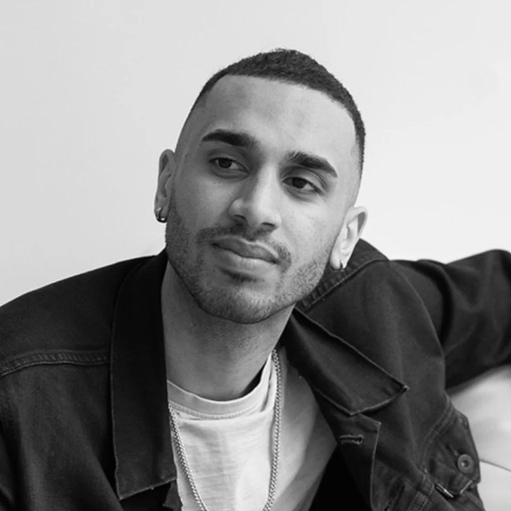 Black and white photo of a young man with short hair, wearing a denim jacket and a white T-shirt, sitting and looking to the side.