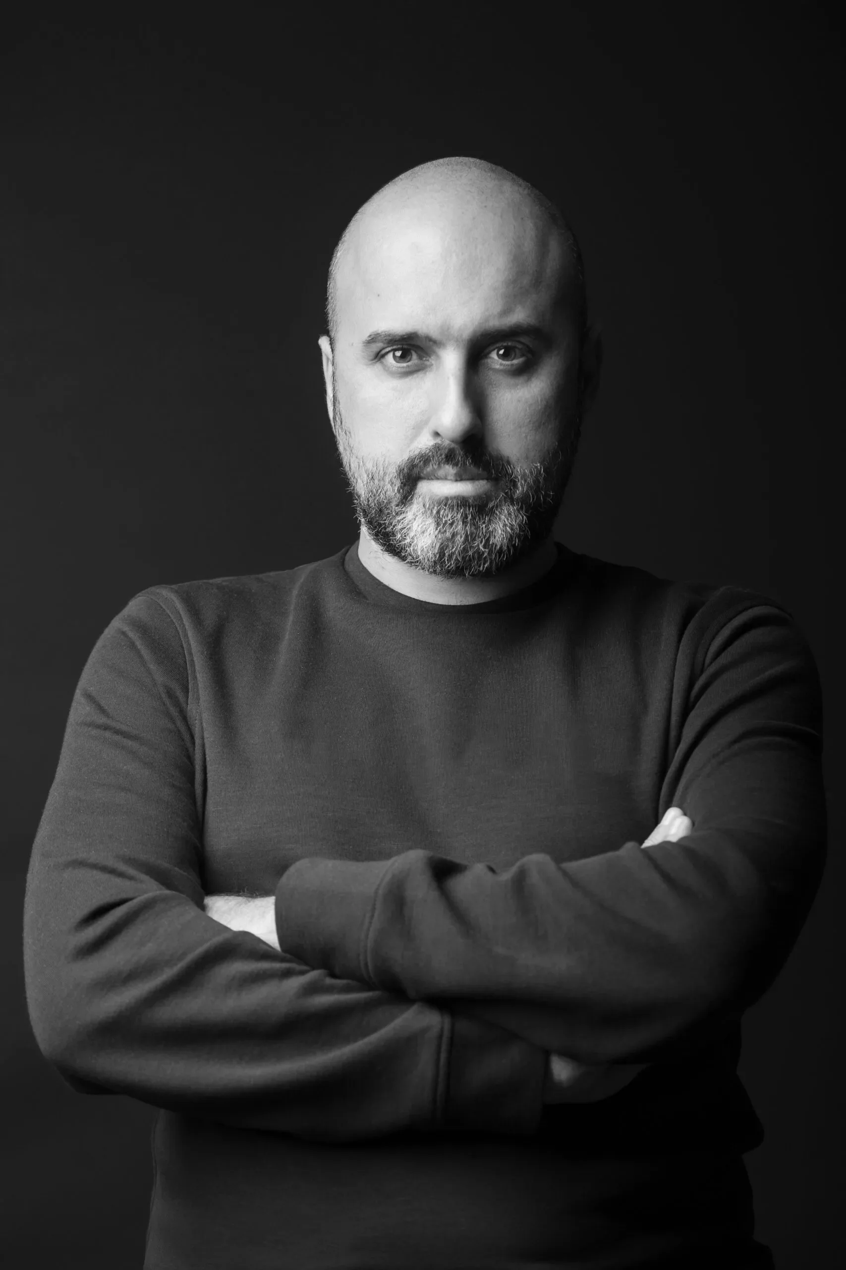 Black and white portrait of a bald man with a beard, arms crossed, wearing a dark long-sleeve shirt, against a plain dark background.