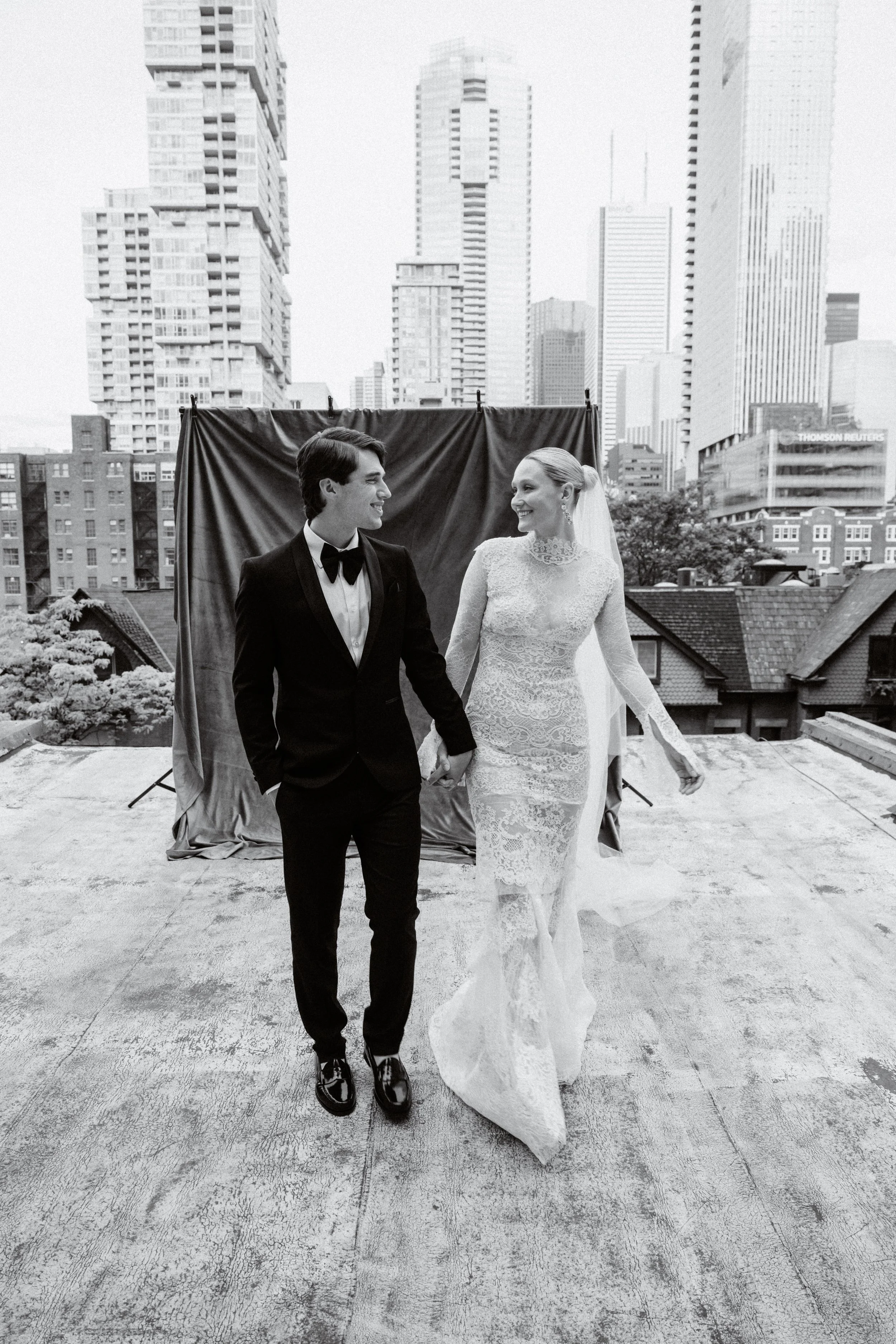 A bride and groom holding hands and smiling at each other on a rooftop with city skyscrapers in the background, black and white photo.