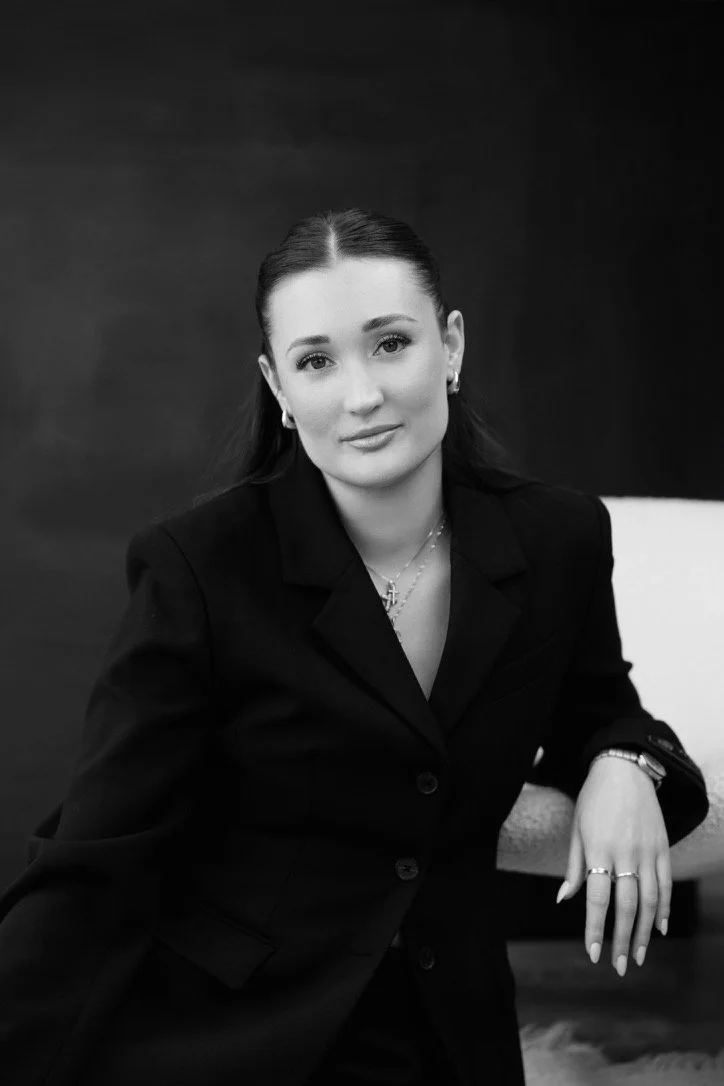 A woman in a formal black suit sitting on a chair, looking at the camera in a black-and-white portrait.