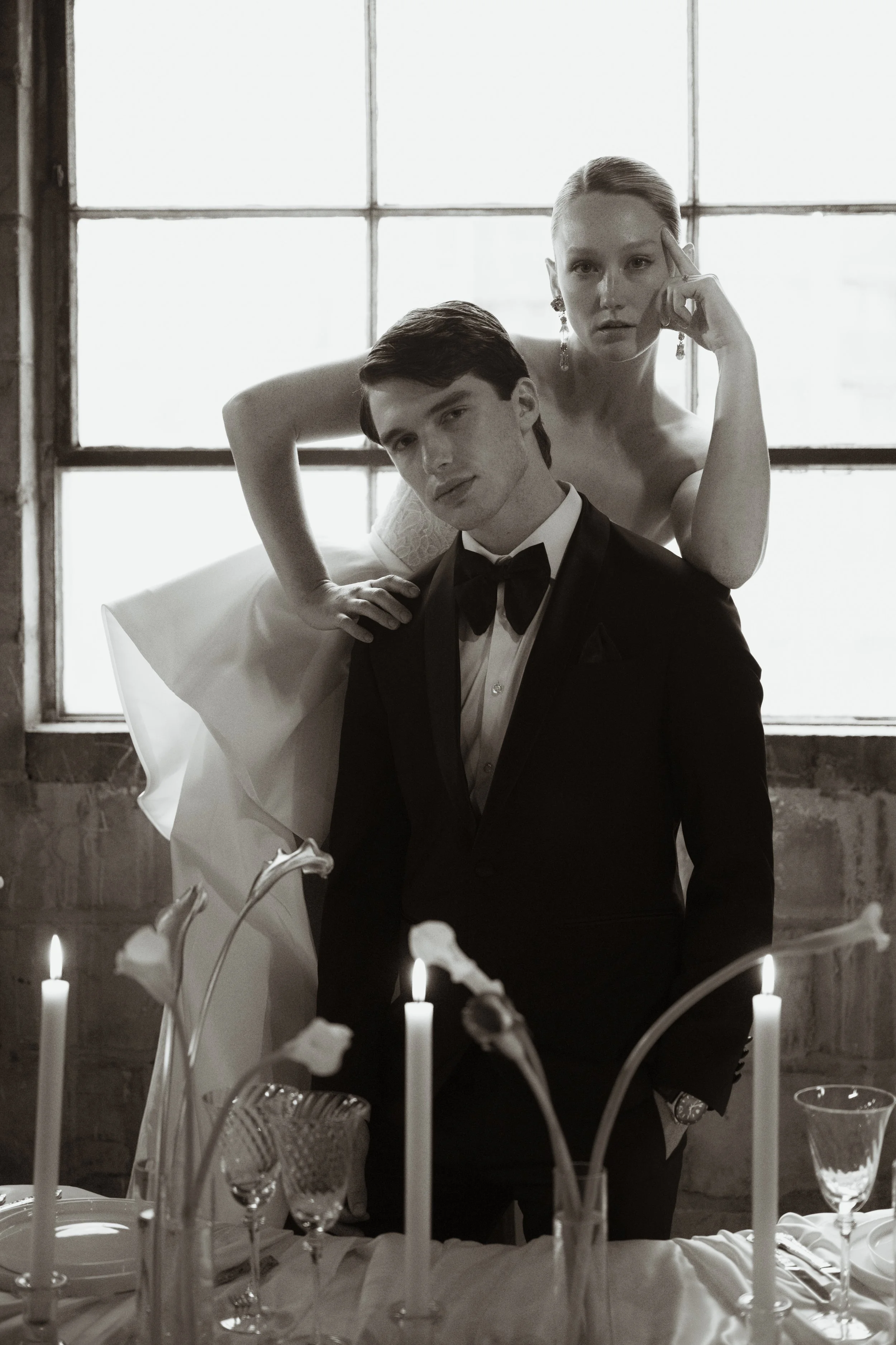 Black and white photo of a man in a tuxedo and a woman in a wedding dress posing behind a dining table with lit candles and glassware, standing in front of a large window.