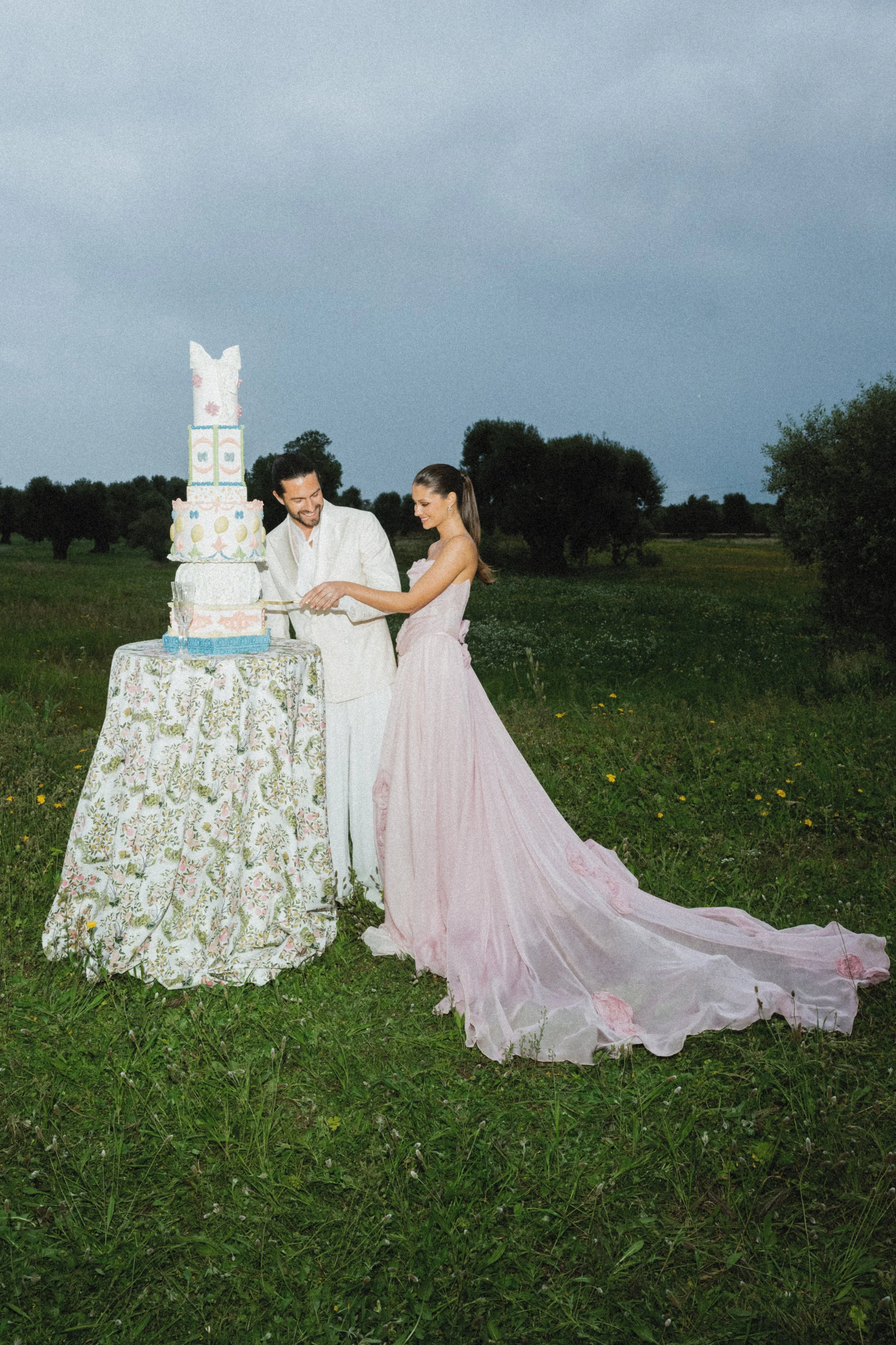 A couple in formal attire cuts a tiered wedding cake outdoors on a grassy field with trees in the background.