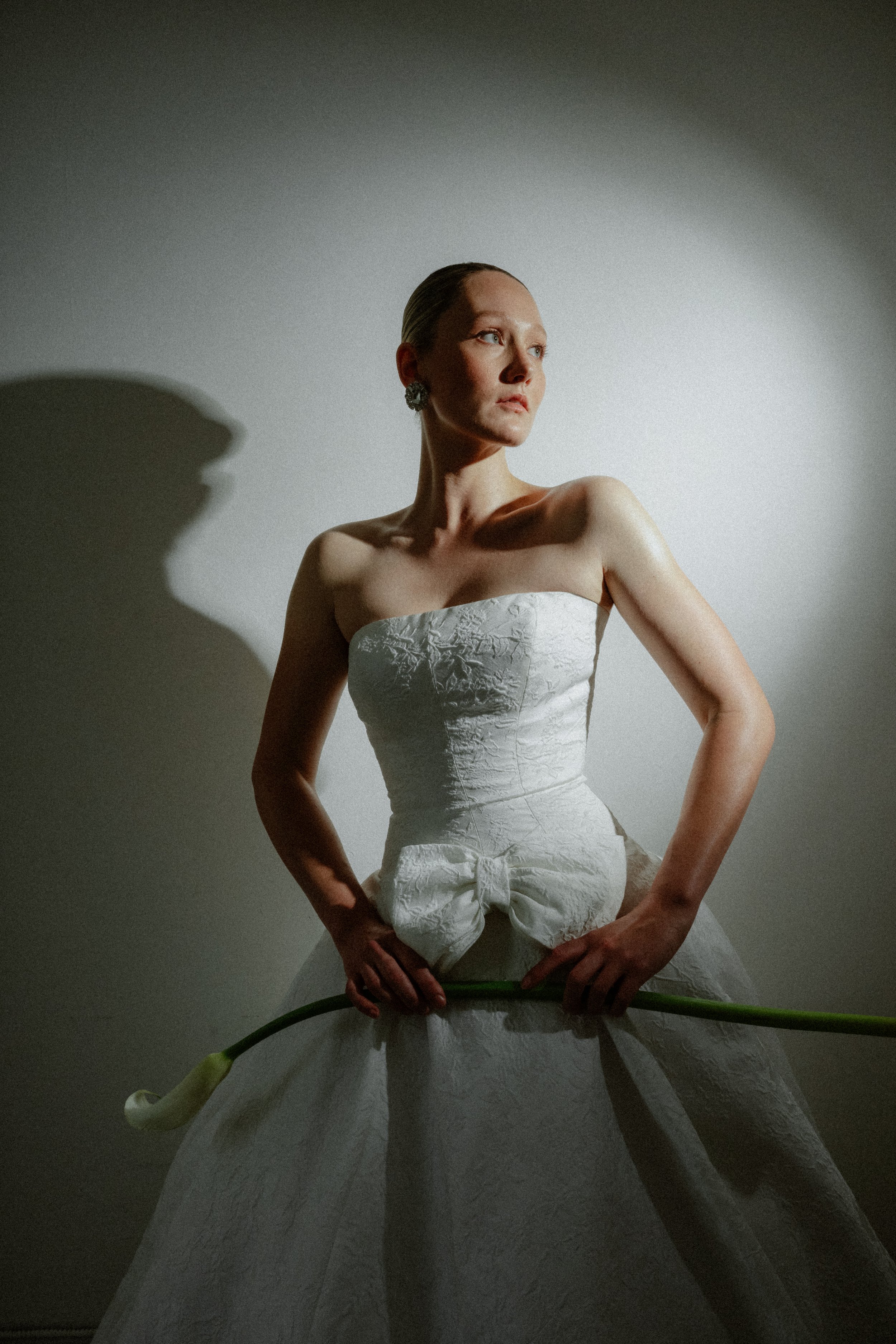 A woman in a strapless wedding dress holding a calla lily flower diagonally across her waist, standing against a plain wall with lighting casting a shadow.