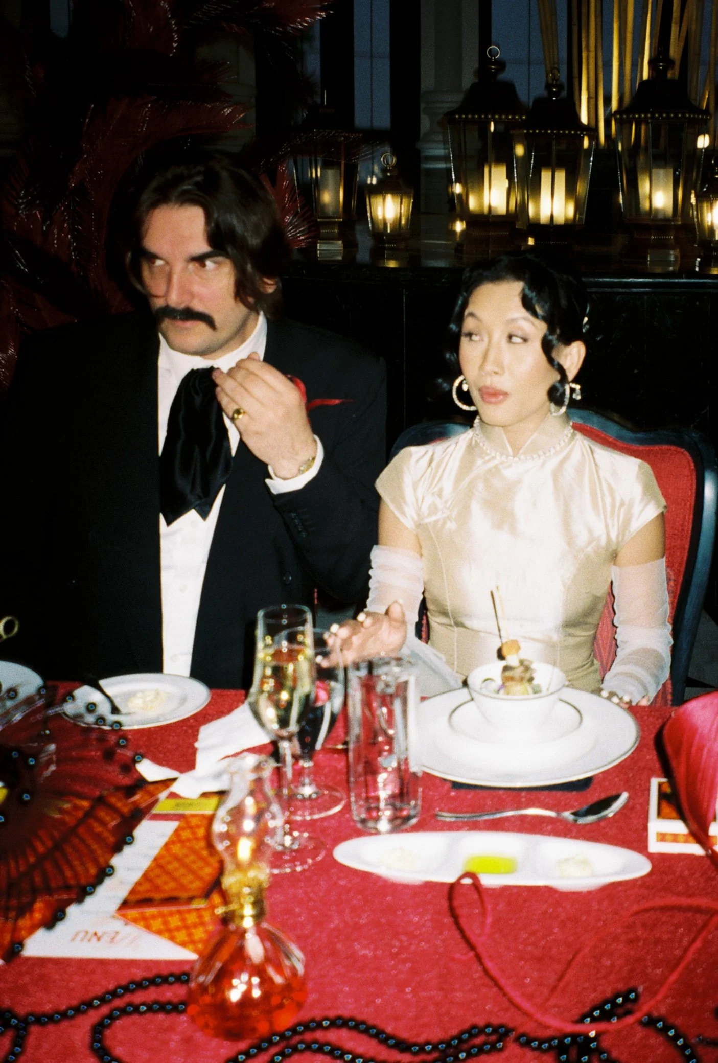 A man with dark hair, a mustache, and dressed in a tuxedo with a white shirt and black bow tie, is sitting at a festive table with a woman who has short black hair styled in vintage waves and is wearing a silky, cream-colored dress. The table is decorated with red tablecloths, glassware, and Mardi Gras beads. The background features candle lanterns on a dark surface and dark curtains, suggesting a formal or themed celebration event.
