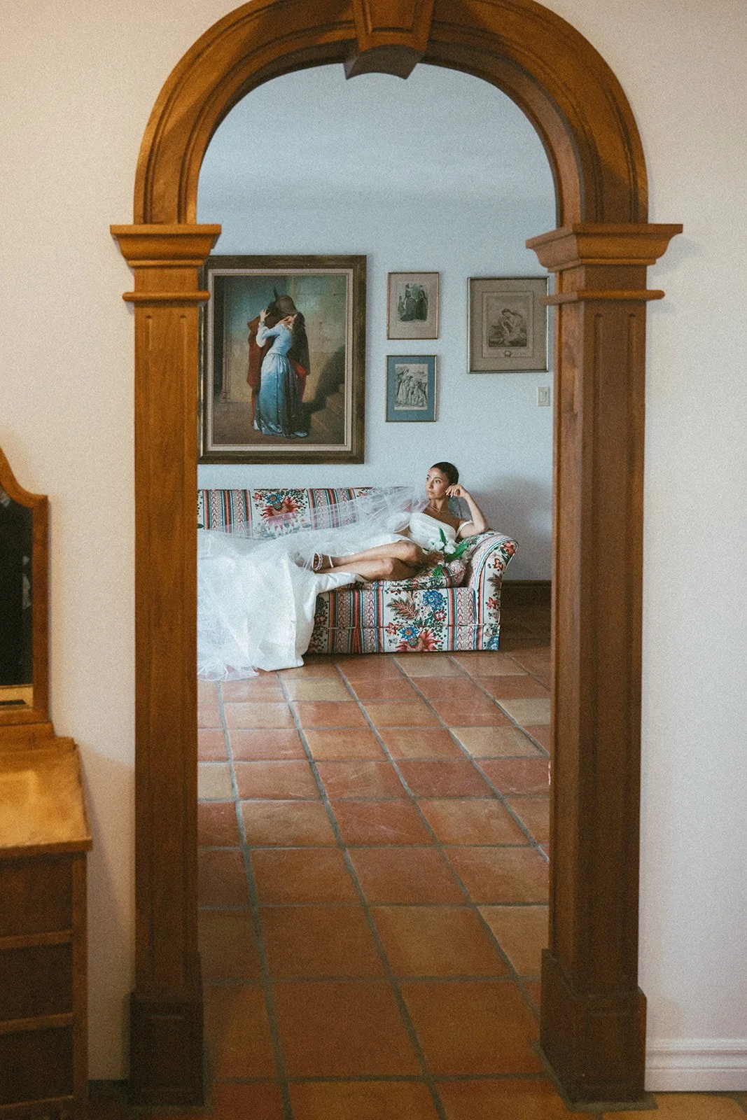 A bride in a wedding dress sitting on a colorful patterned sofa inside a room, viewed through a wooden archway, with framed artwork on the wall behind her.