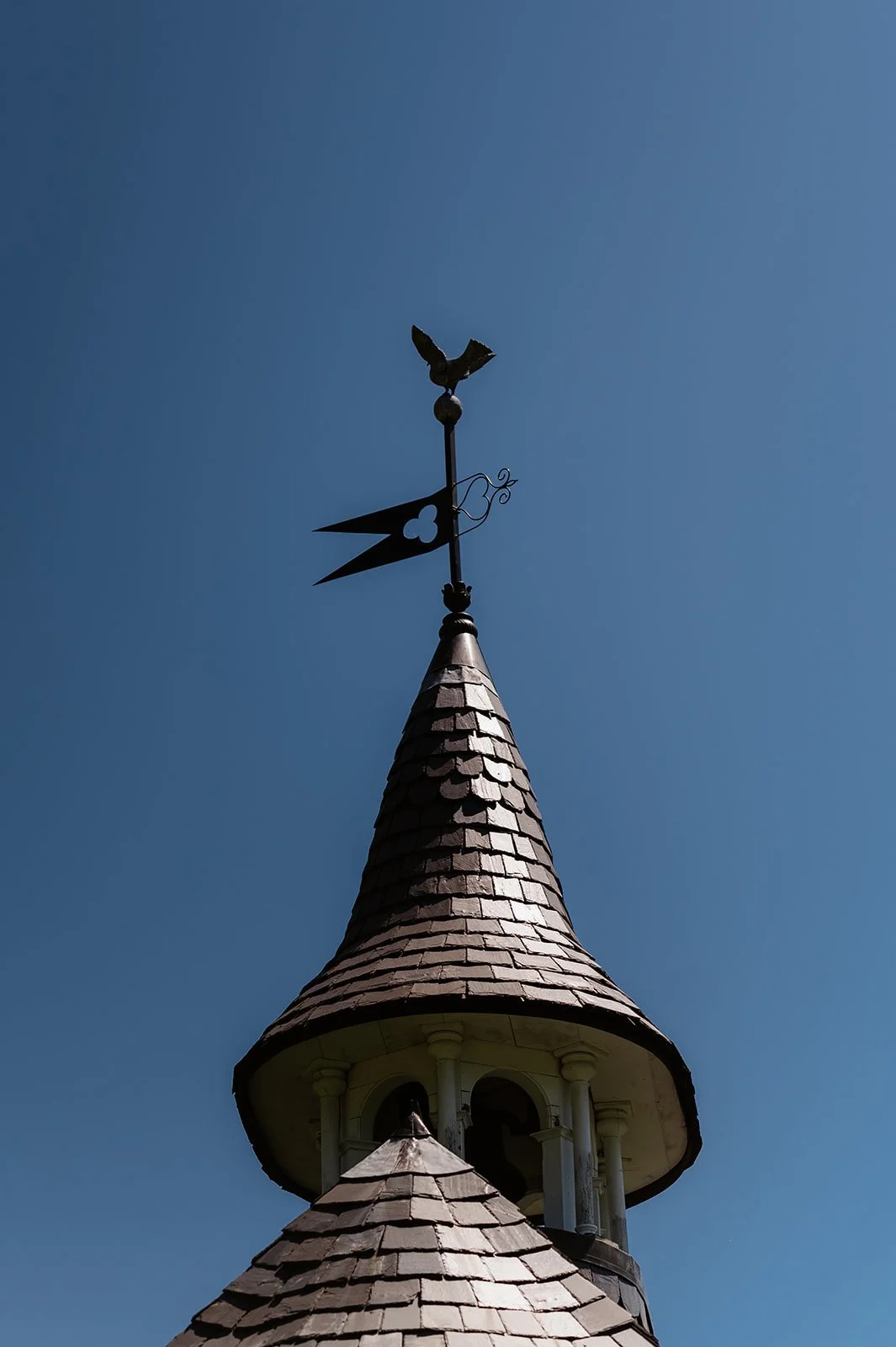 A weather vane with a bird figure on top mounted on the spire of a church steeple against a clear blue sky.