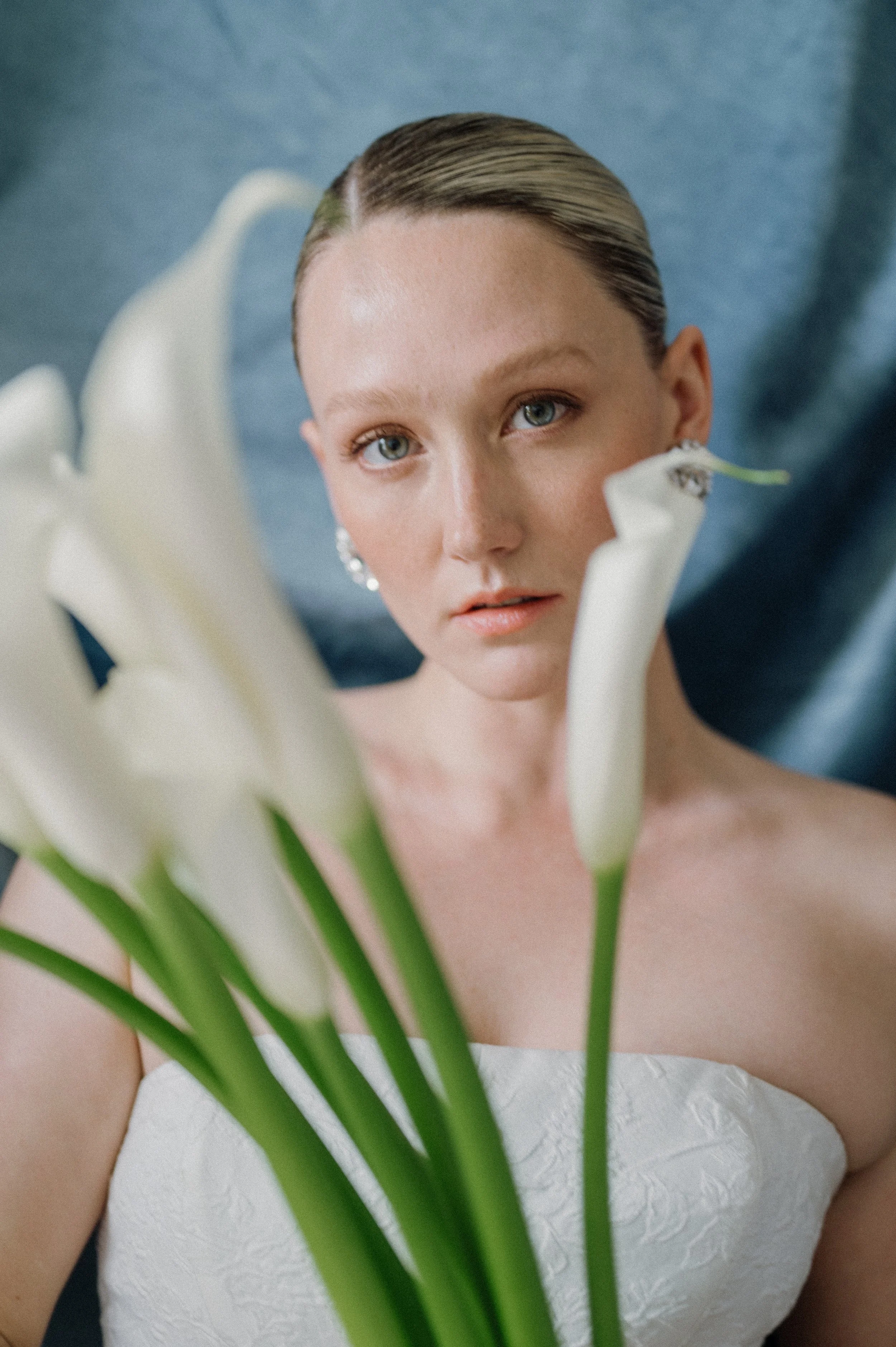A woman with blue eyes and blonde hair styled in a sleek, wet look, wearing pearl earrings and a strapless white dress, poses behind a bouquet of white calla lilies in front of a dark blue background.
