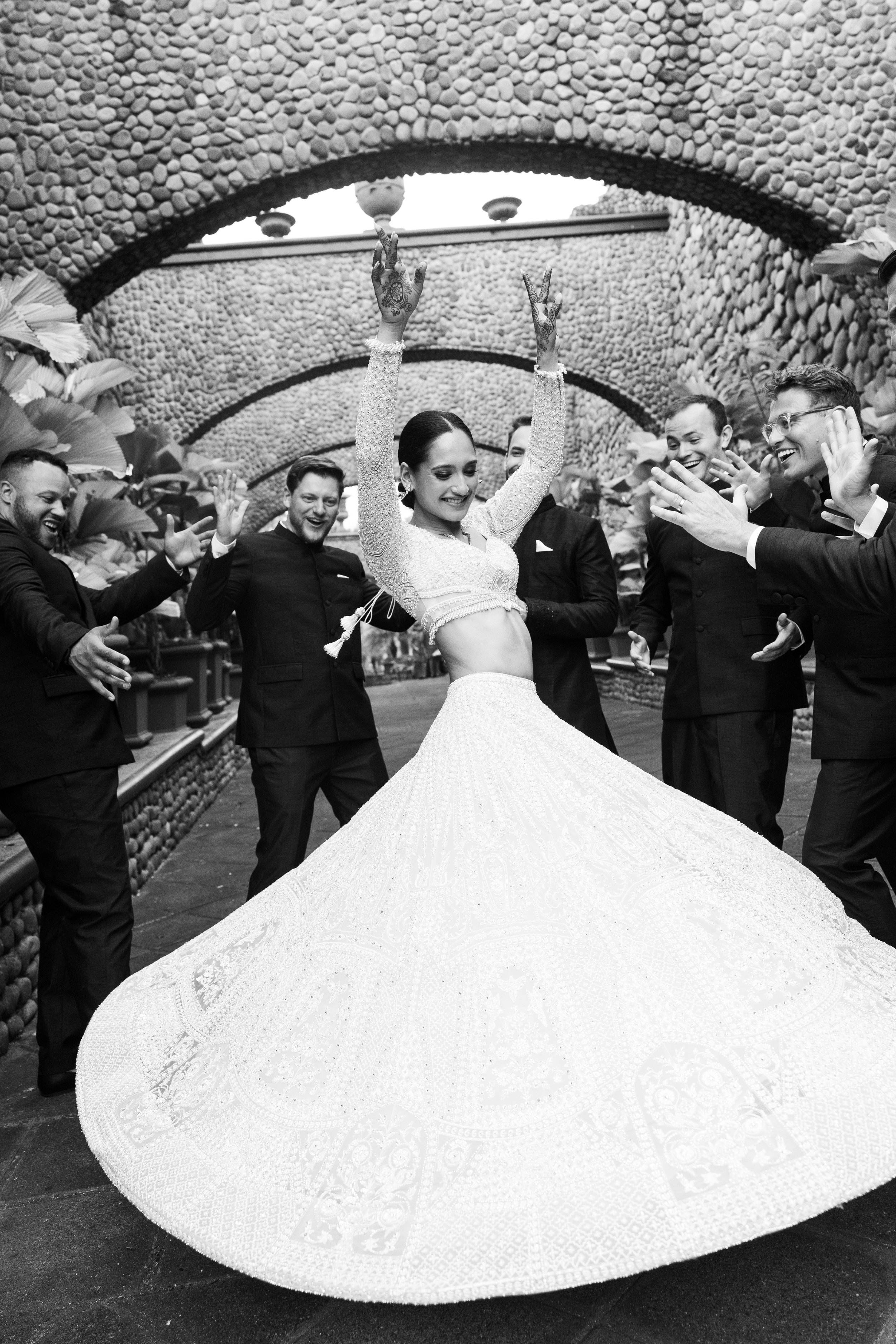 A woman in a traditional Indian wedding saree dancing with a group of men in formal suits at a wedding celebration under arched stone structures.