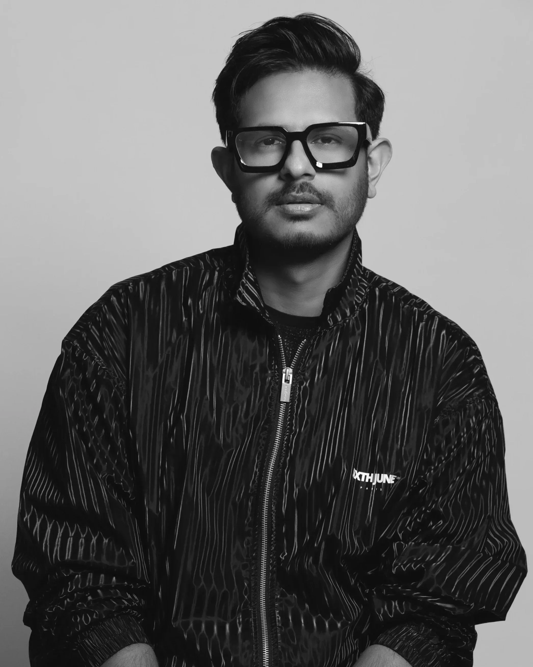 Black and white portrait of a young man with styled hair, wearing large black glasses, a patterned zip-up jacket, and a serious expression.