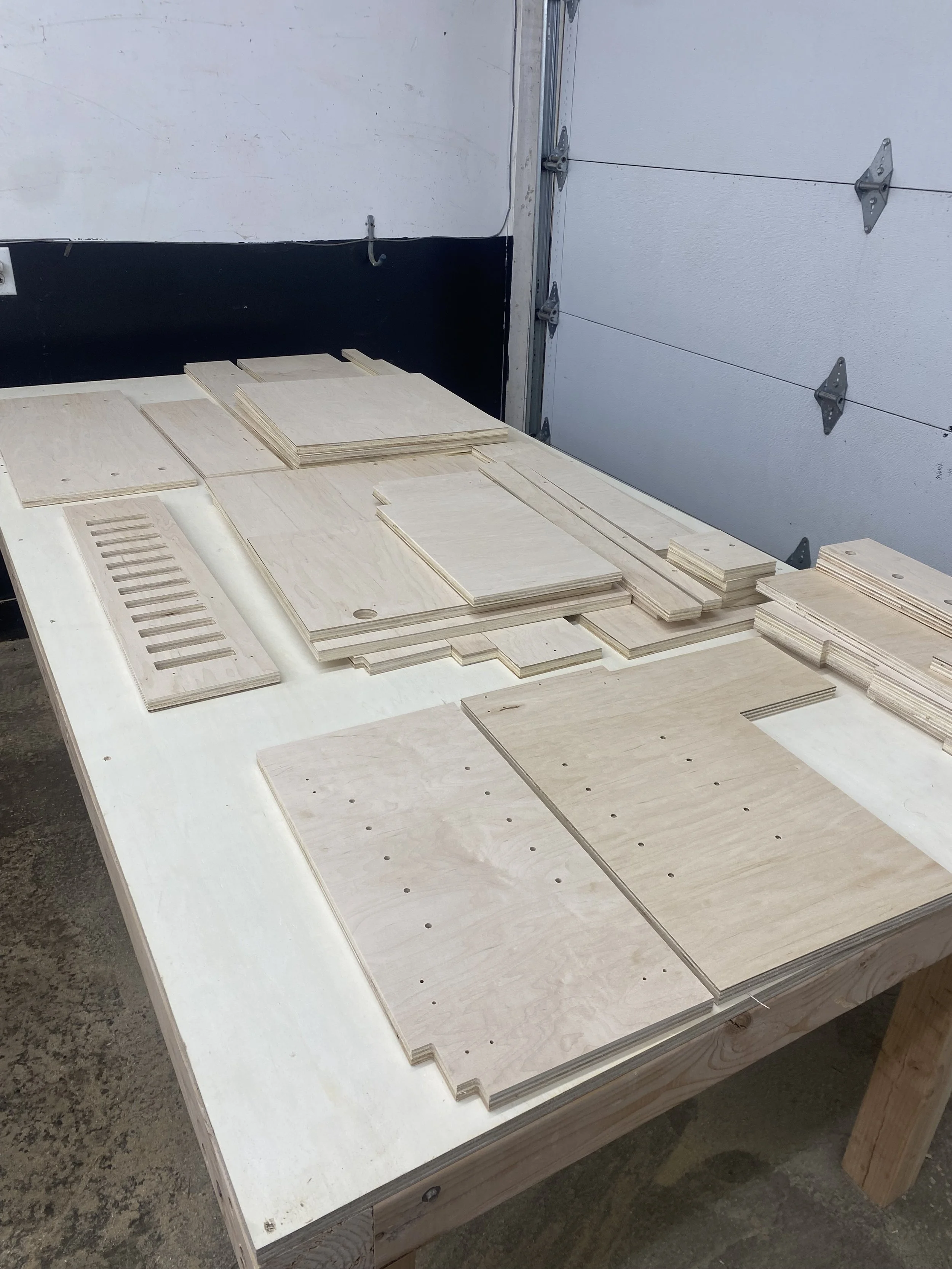 Sheets of plywood with drilled holes are arranged on a white work table in a woodworking or construction workspace.