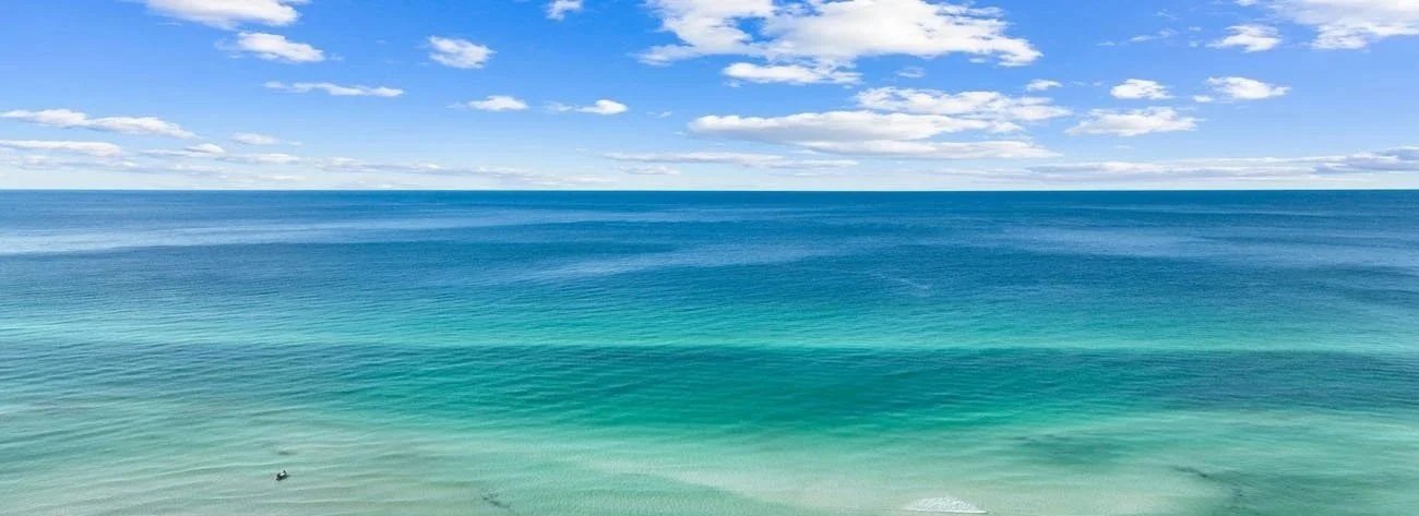 A panoramic view of the ocean with a sailboat in the distance under a partly cloudy sky.