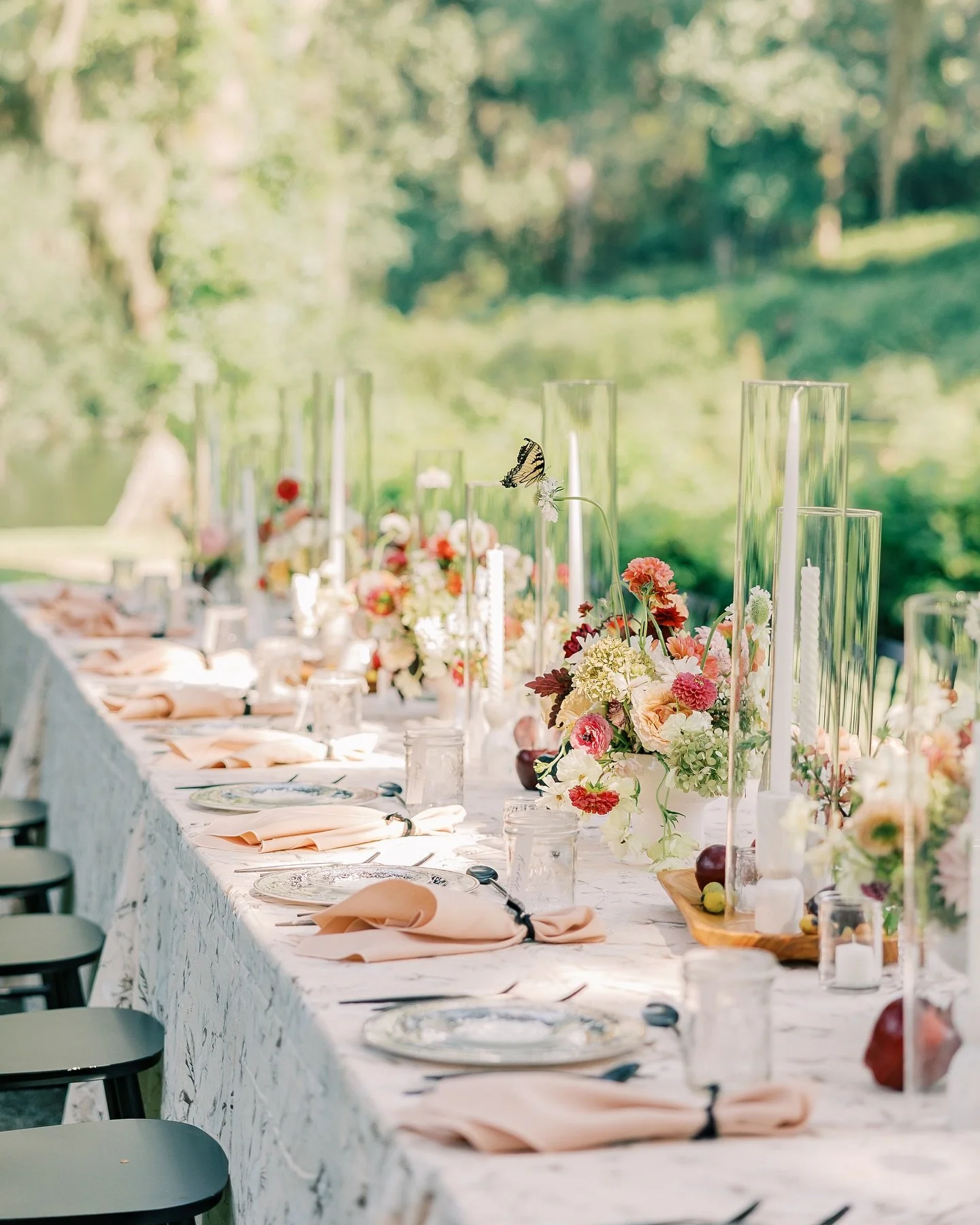 Outdoor dining table decorated with floral arrangements, candles, and glass candle holders, set for a special event.