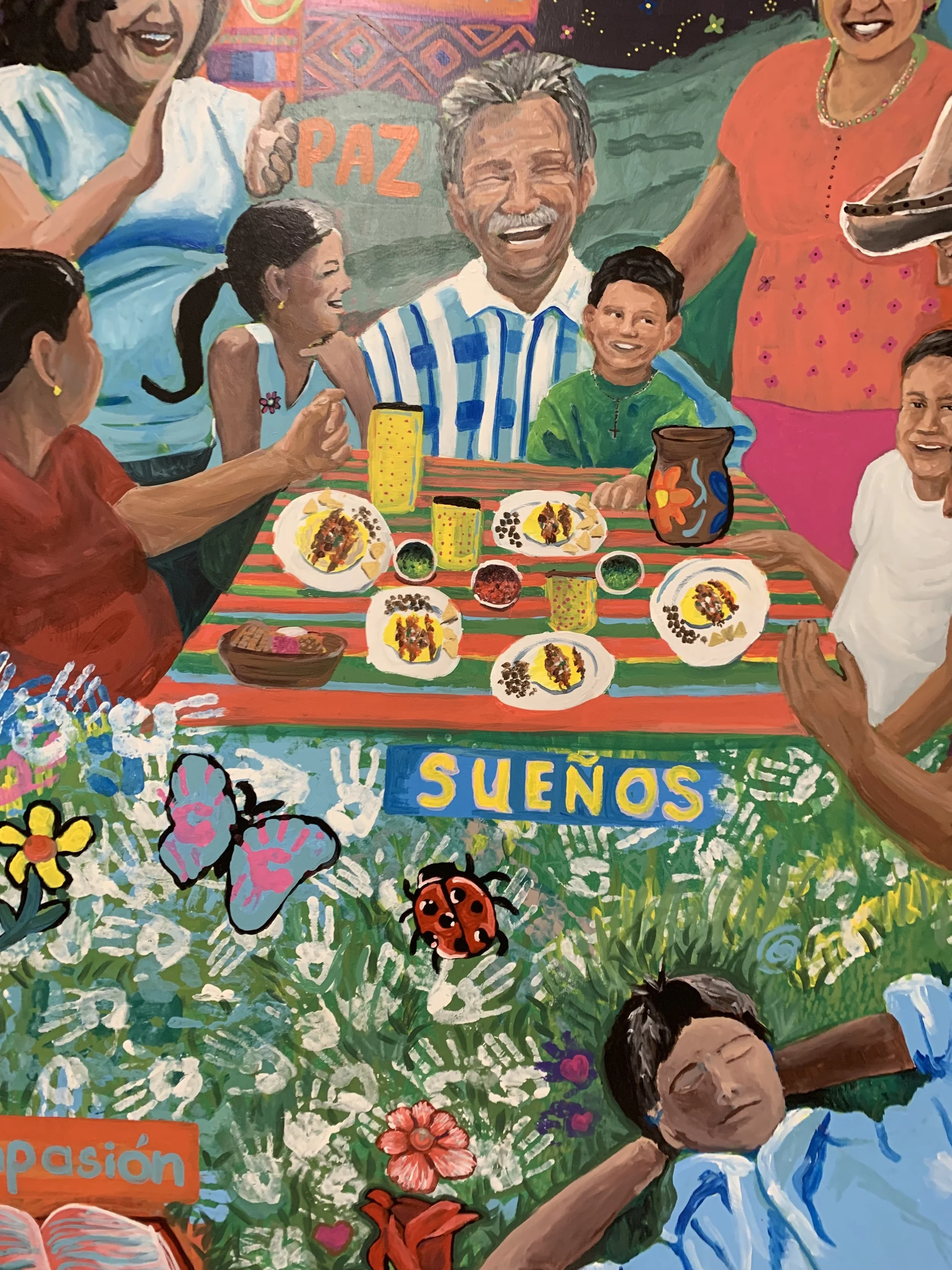 A family of eight gathered smiling and laughing around a colorful table of tacos and drinks. In the bottom right hand corner, a boy lays in the grass as if daydreaming.
