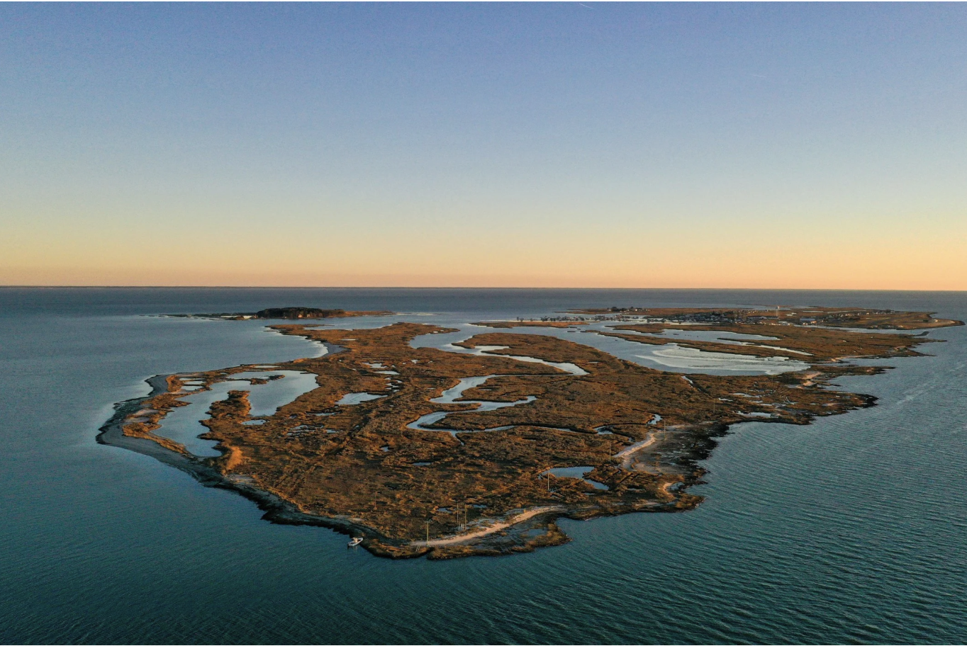 An aerial view of a coastal wetlands area with numerous small water channels, surrounded by land with sparse vegetation, under a clear sky at sunset.