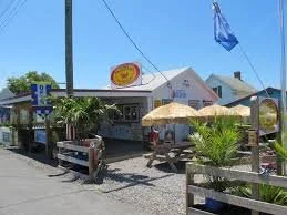 A small restaurant with a white exterior, yellow umbrella, and outdoor seating, situated along a street with a palm tree and a blue flag.