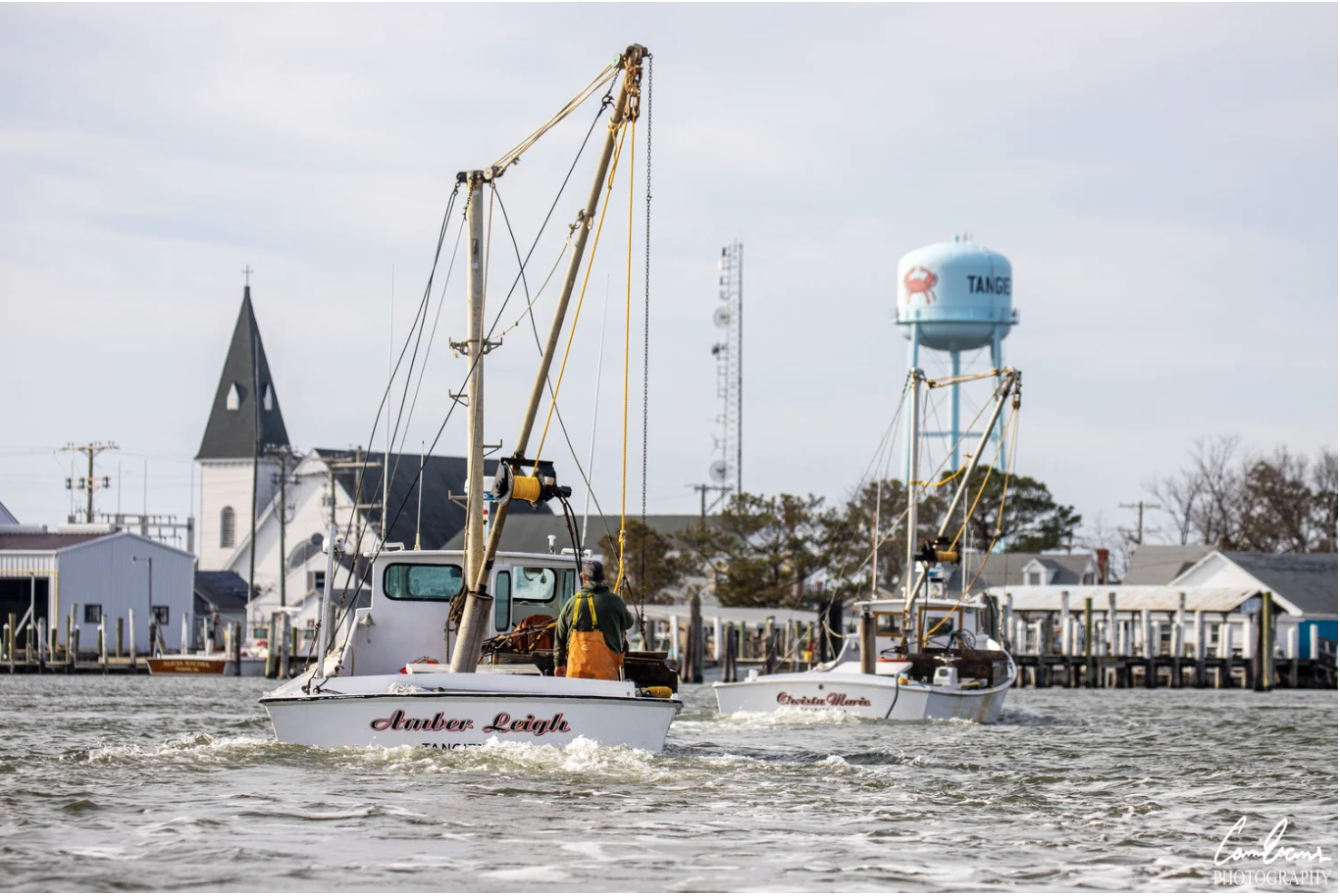 Two small sailboats on the water near a coastal town with a church, houses, and a water tower in the background.