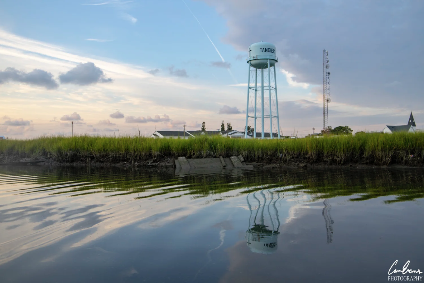 A water tower with 'TANGIER' written on it, reflecting in a body of water, with a partly cloudy sky and some buildings in the background.